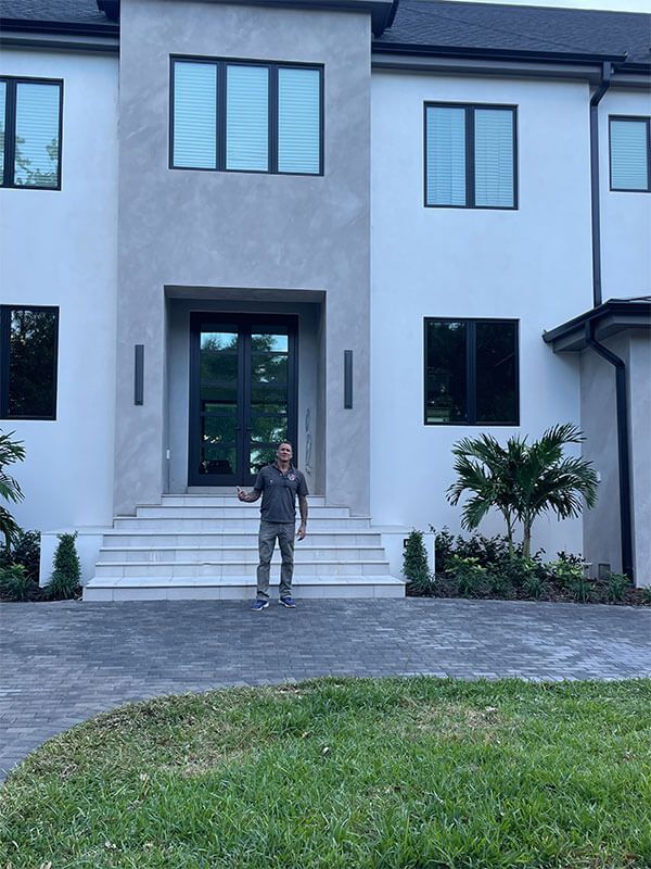 A person stands on the front steps of a modern white two-story house with a grey stone facade and dark-framed windows.