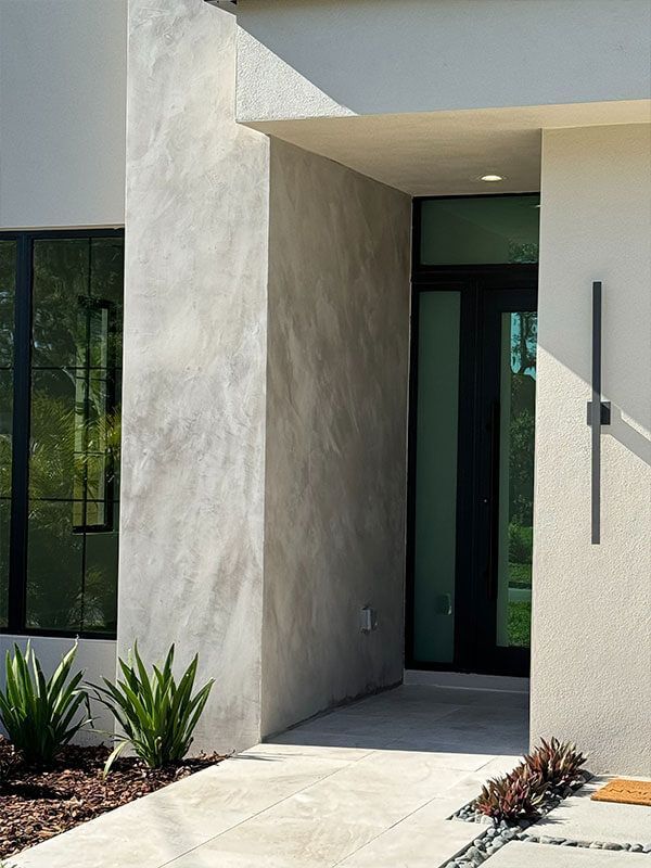 Modern house entrance with a black door, textured gray accent wall, and a vertical black light fixture on a white wall.