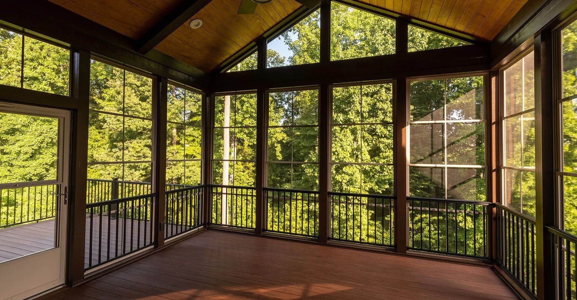 Sunlit screened porch with dark railings, wooden floor, and forest views from large windows