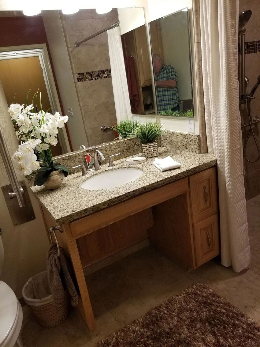 Bathroom vanity with granite countertop, large mirror, sink, and white flowers beside a brown rug.