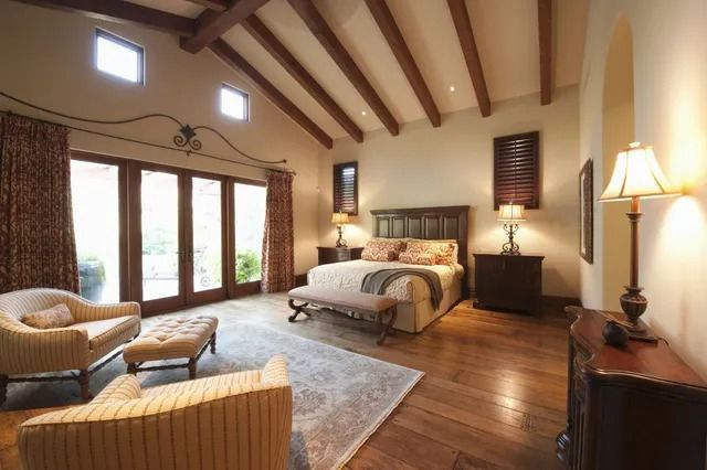 Spacious rustic bedroom with exposed beams, king bed, lounge chair, and large windows with curtains