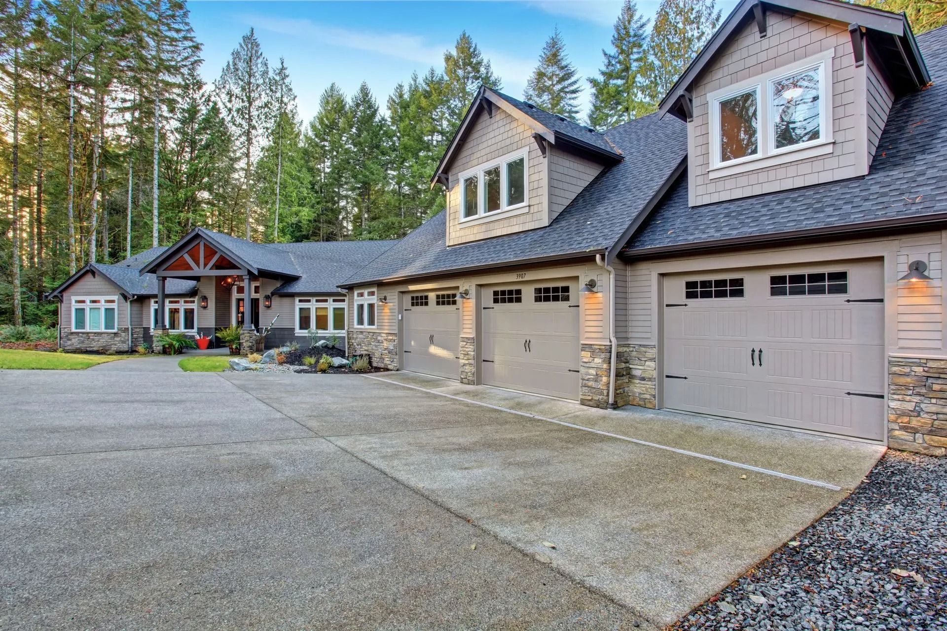 Large modern house with stone garage, gravel driveway, and tall pine trees in front