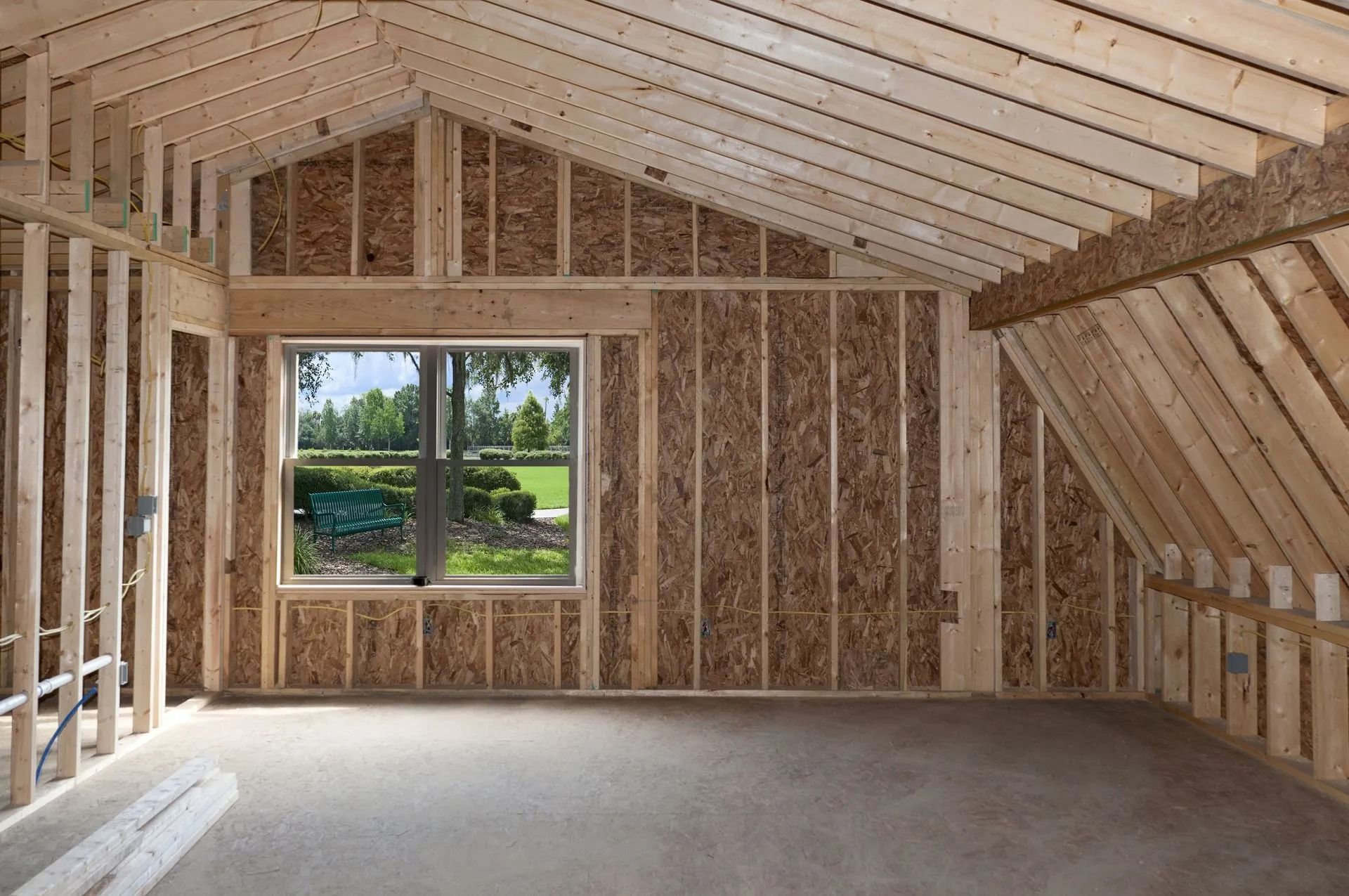 Unfinished attic room with exposed framing, insulation, and a window overlooking greenery.