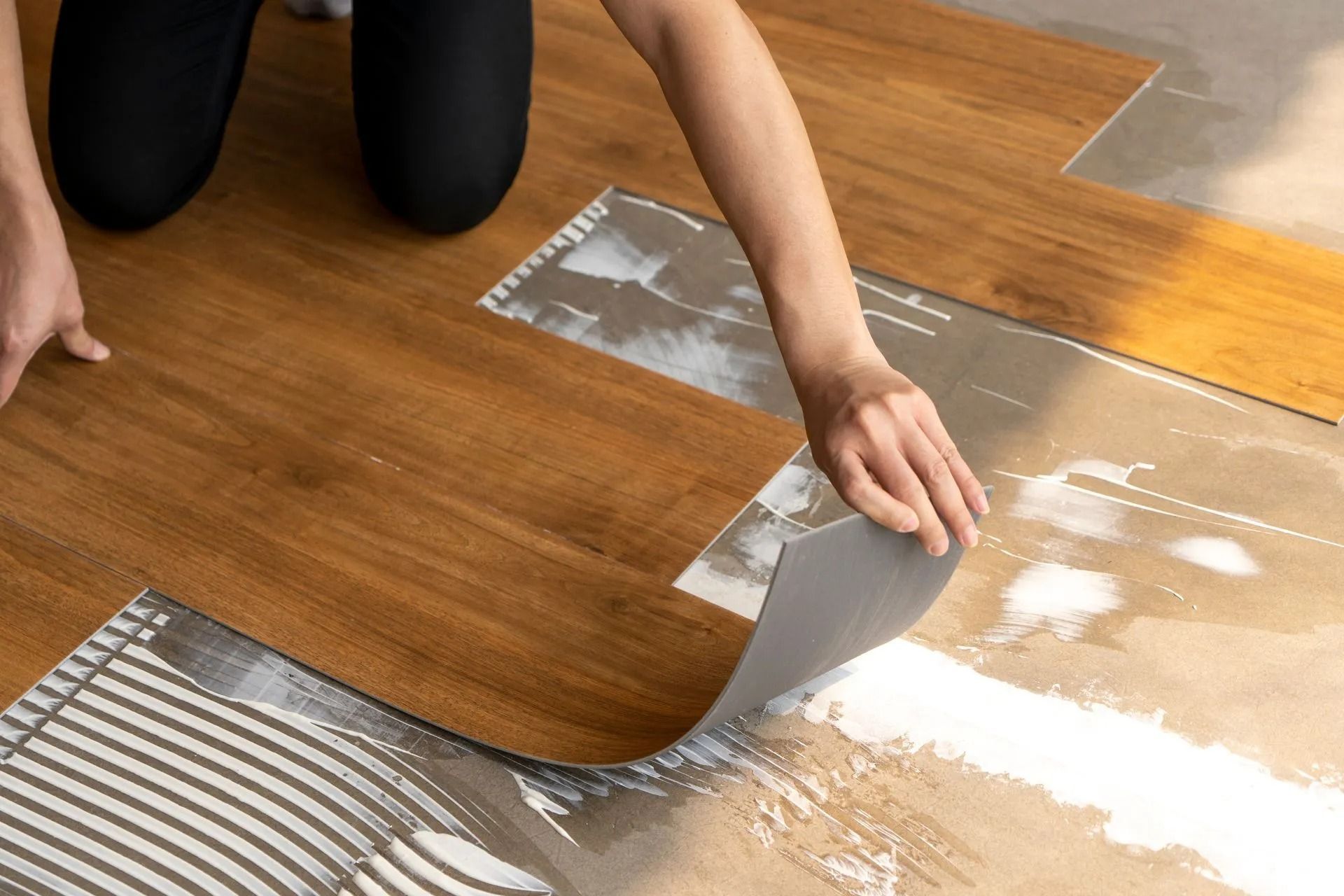 Person peels back floor tile or adhesive sheet to reveal wood flooring beneath