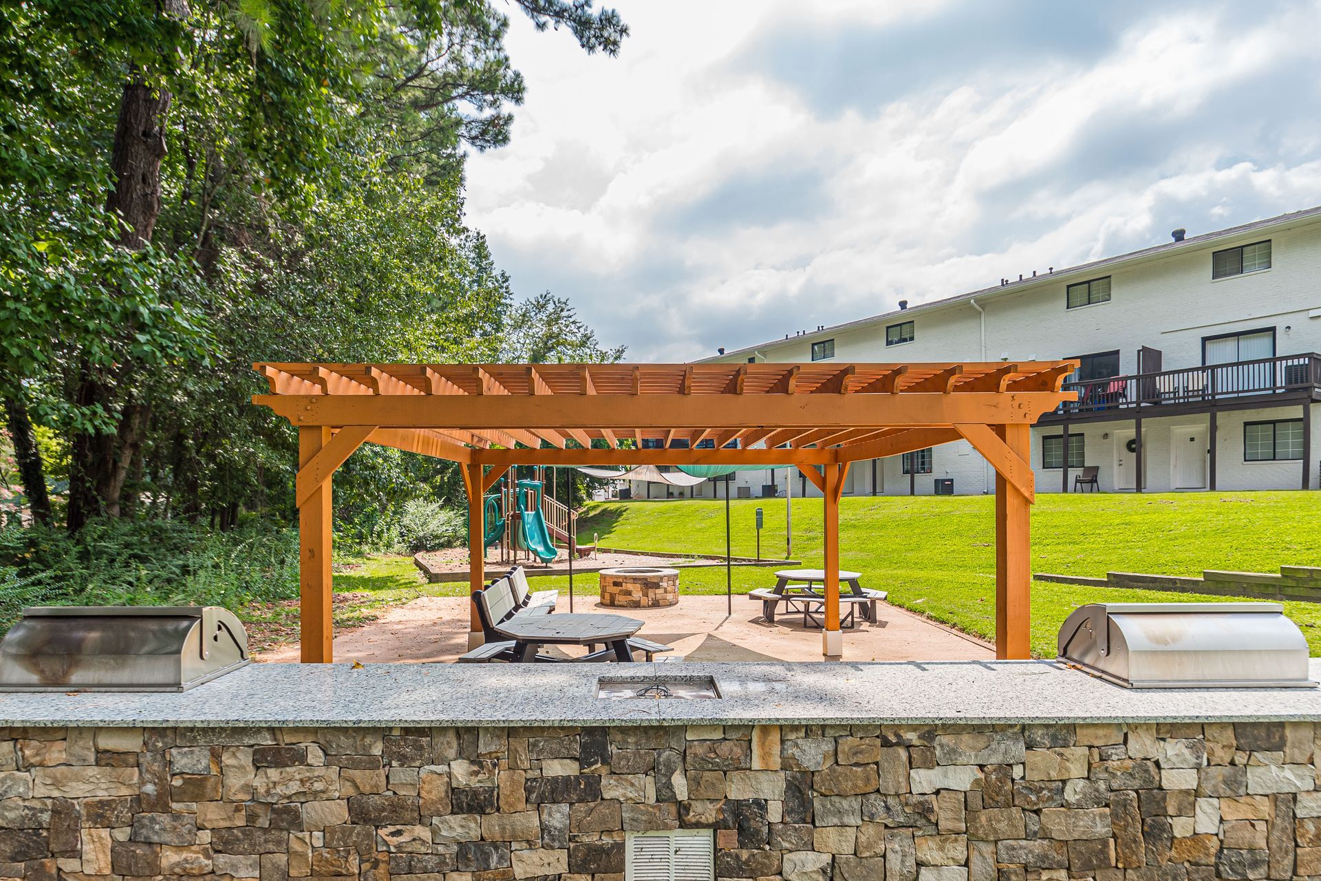Wooden pergola over a picnic area in a sunny park with stone seating and trees nearby