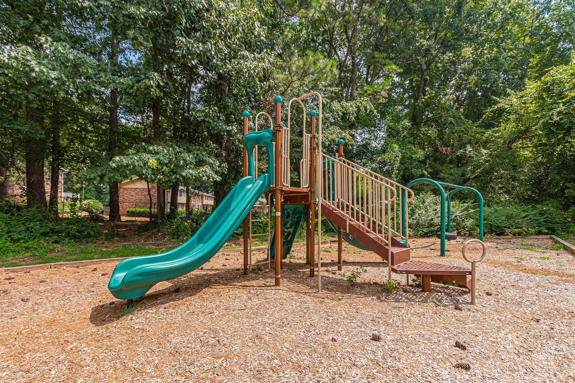 Empty playground with a teal slide, steps, and wood chips under trees.