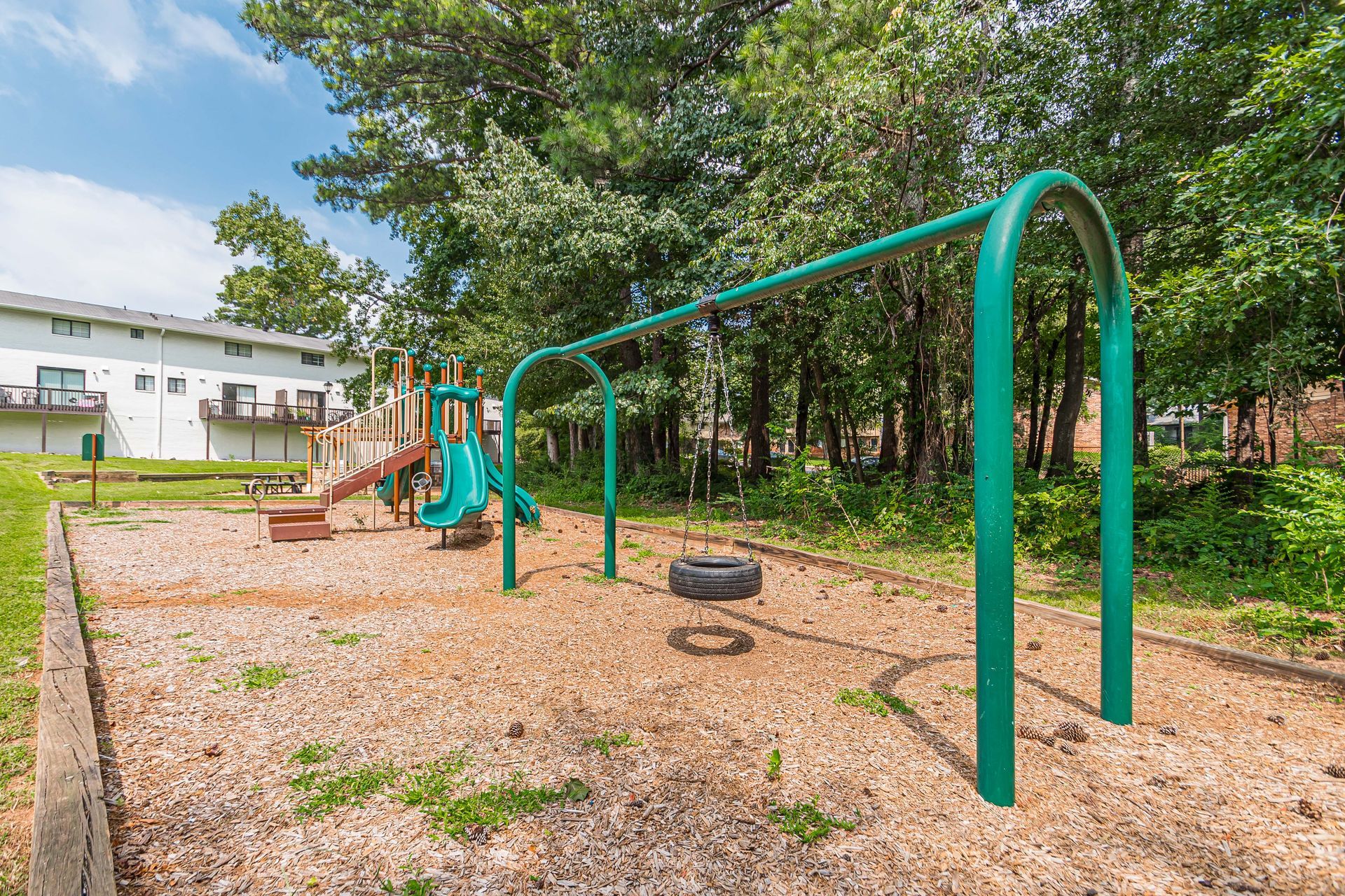 Empty playground with green monkey bars, swings, and rubber tire on wood-chip ground near trees and buildings