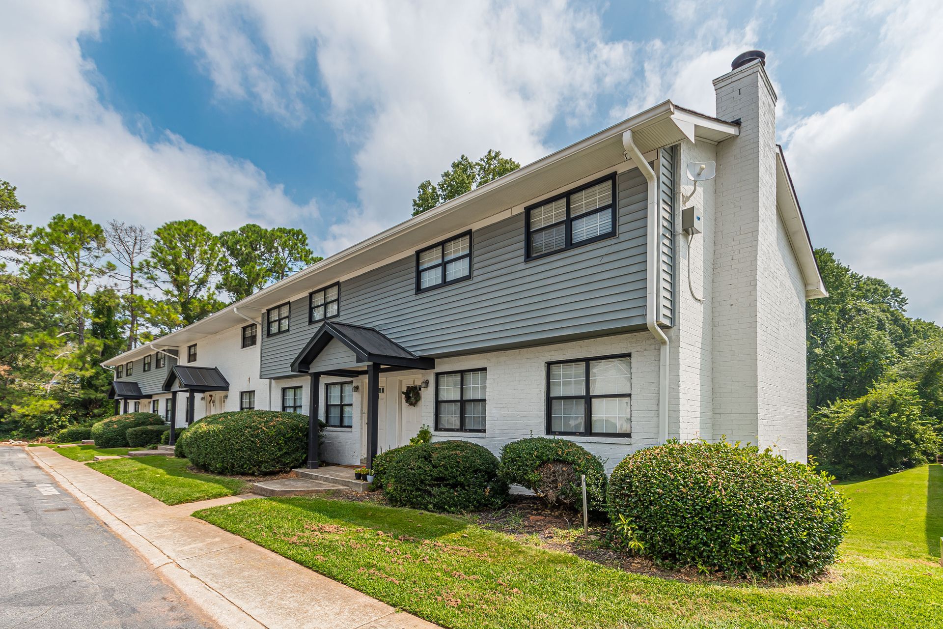 Two-story gray apartment building with white trim, shrubs, and a sidewalk under a partly cloudy sky