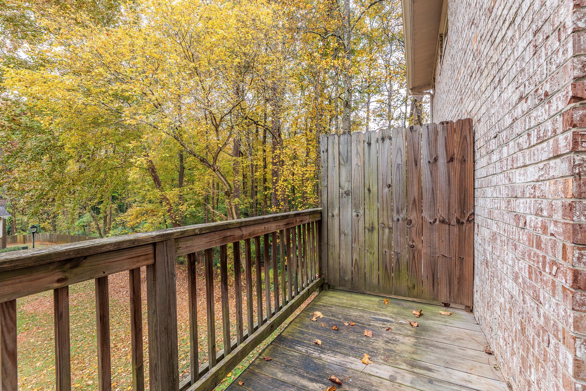 Wooden balcony beside a brick wall, overlooking autumn trees and a backyard fence.