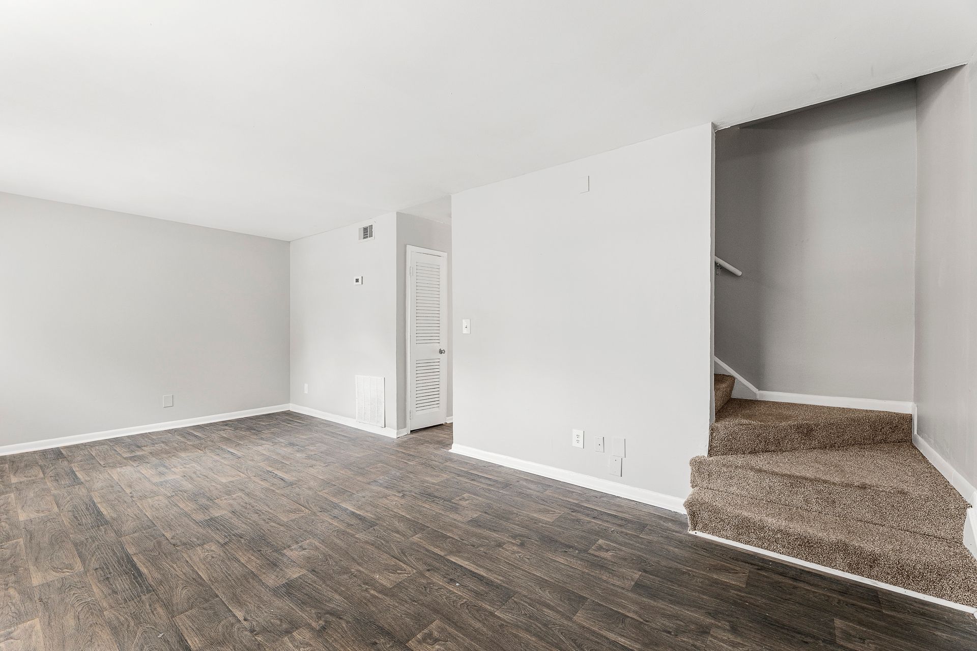 Empty living room with gray wood floors, white walls, and a carpeted staircase opening on the right
