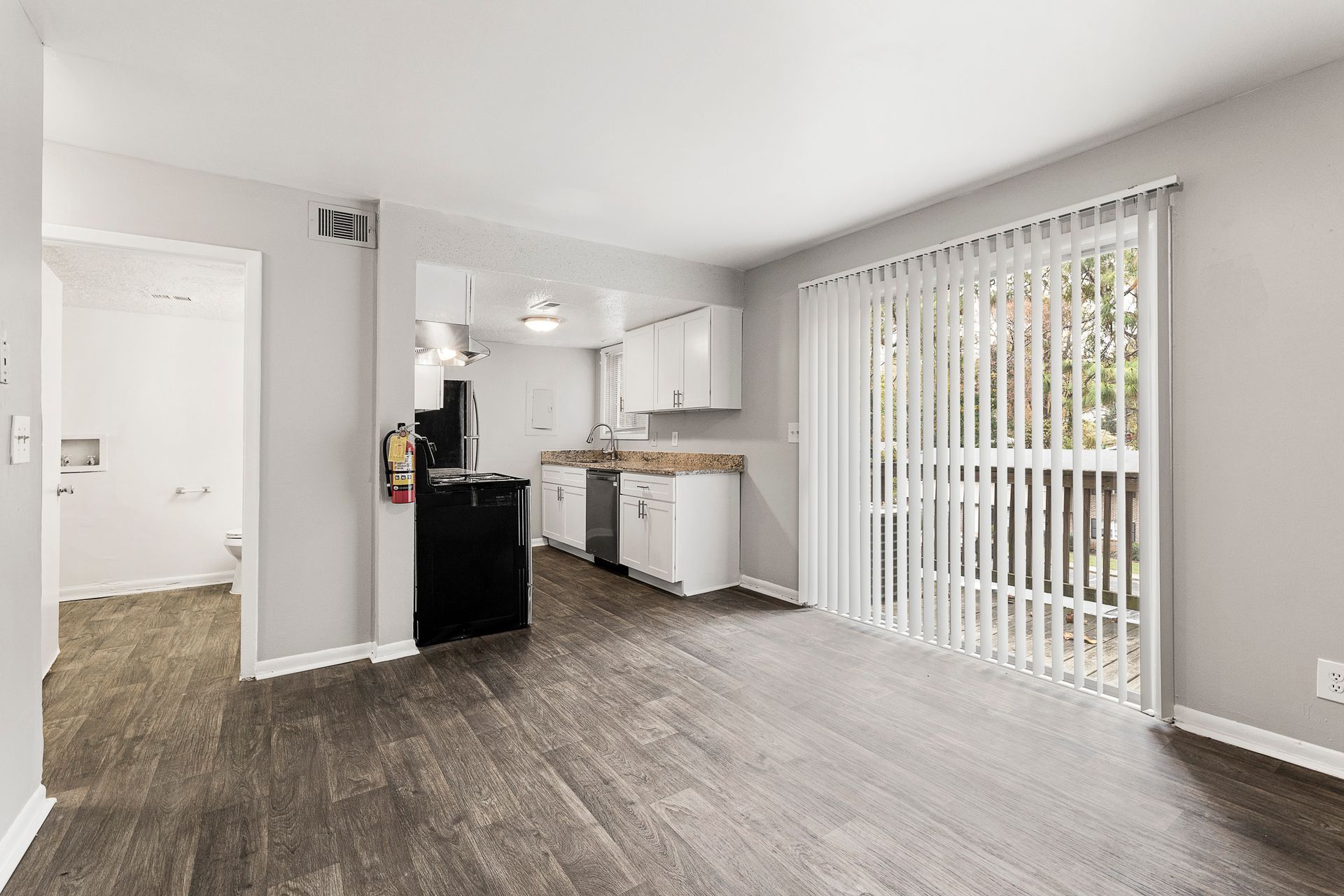 Empty living room with gray wood floors, open kitchen, and sliding glass door with vertical blinds