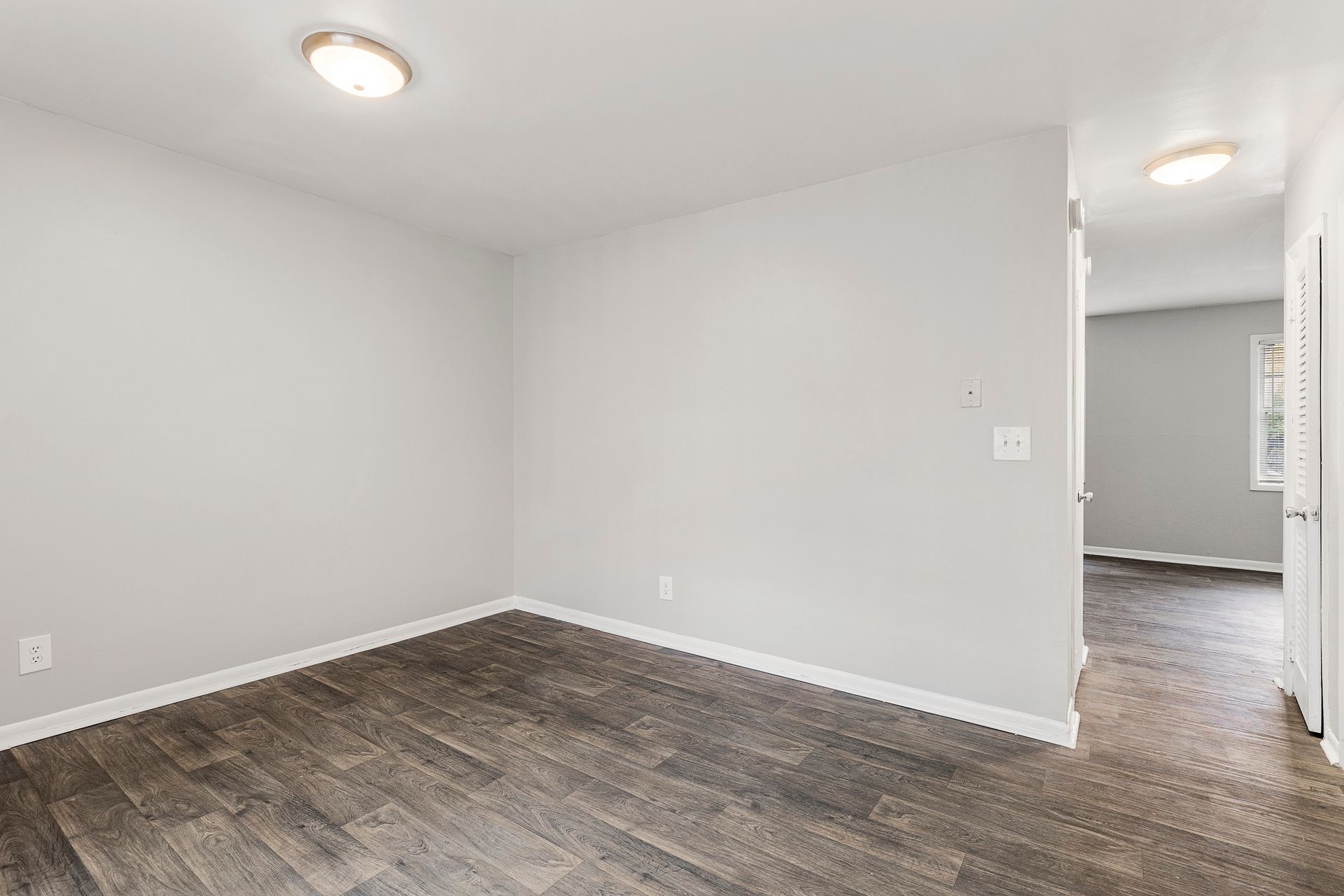 Empty gray room with dark wood flooring and an open doorway to a lit adjacent room
