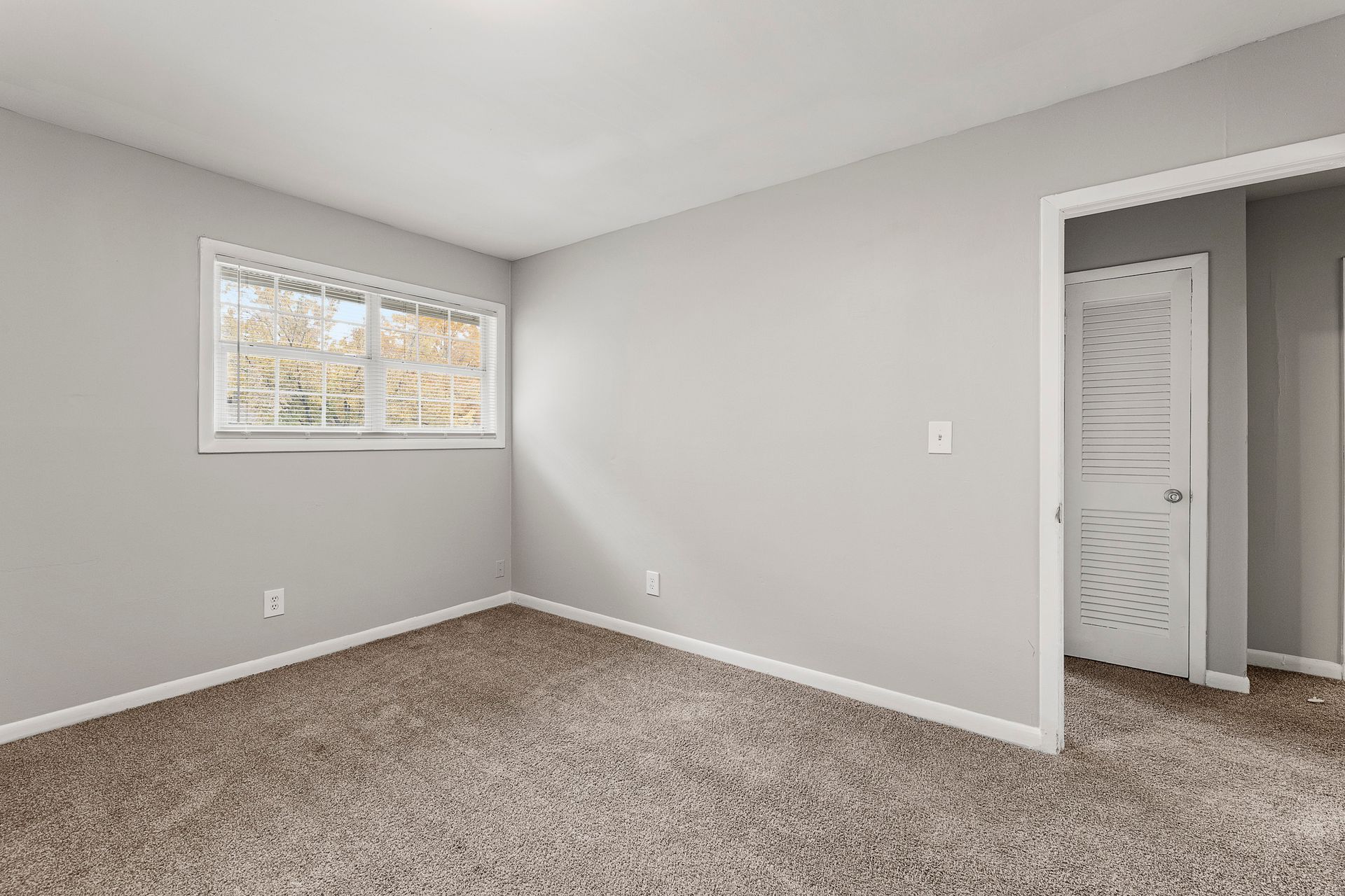 Empty gray carpeted bedroom with a window and open closet doorway