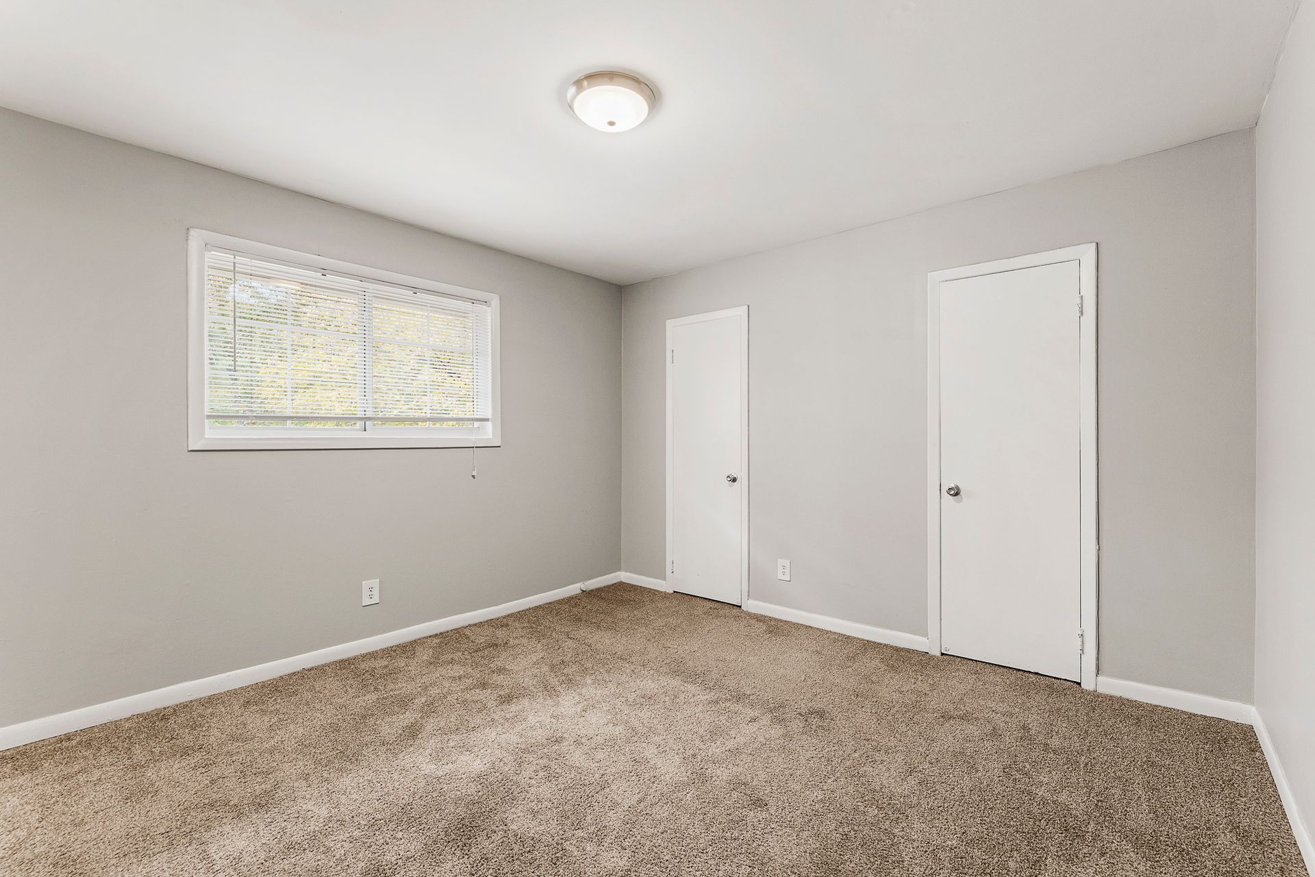 Empty carpeted bedroom with gray walls, white trim, two doors, and a window with blinds.