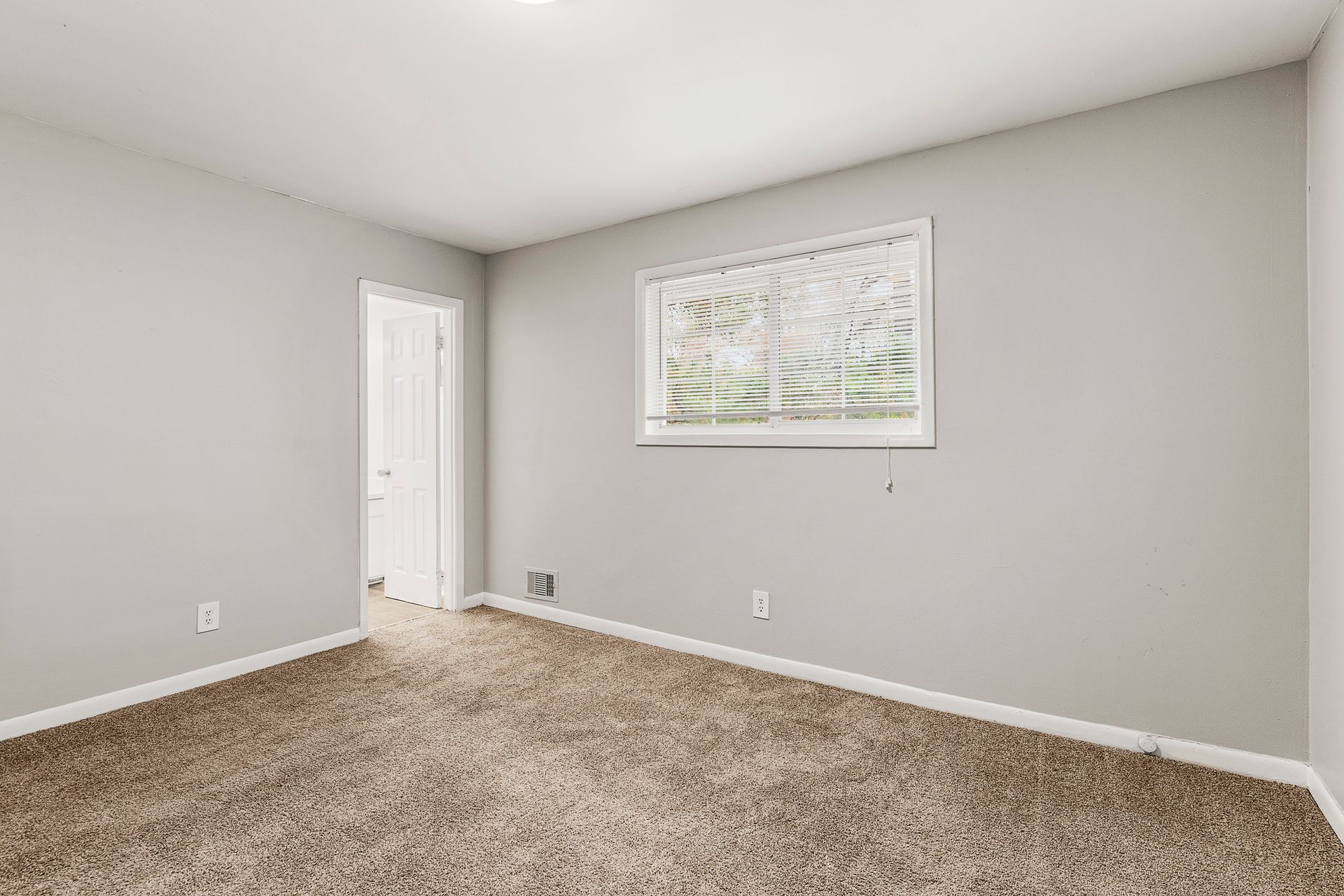 Empty beige bedroom with carpet, gray walls, and a window with blinds.