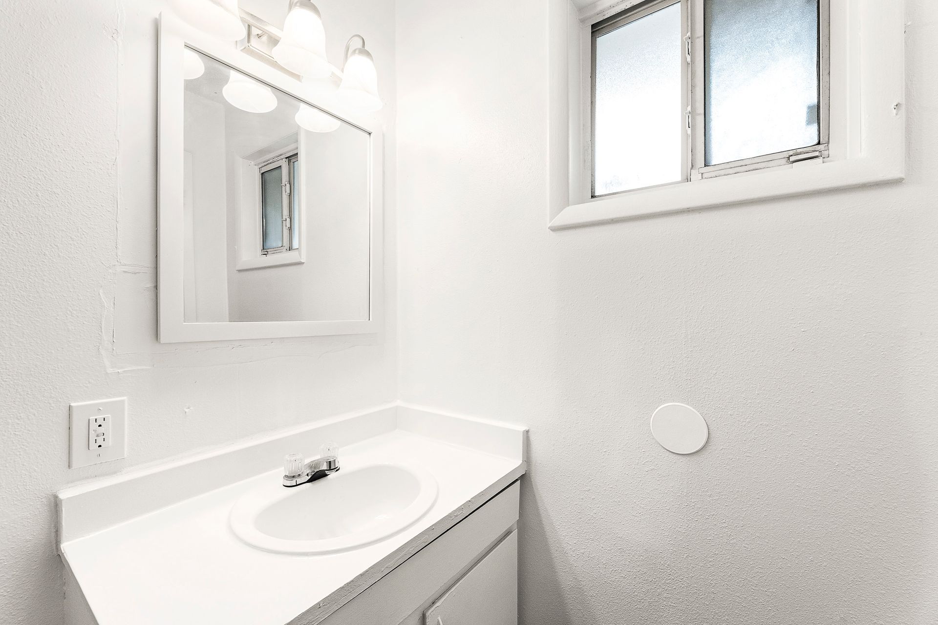 Bright white bathroom corner with vanity sink, mirror, light fixture, and small frosted window.