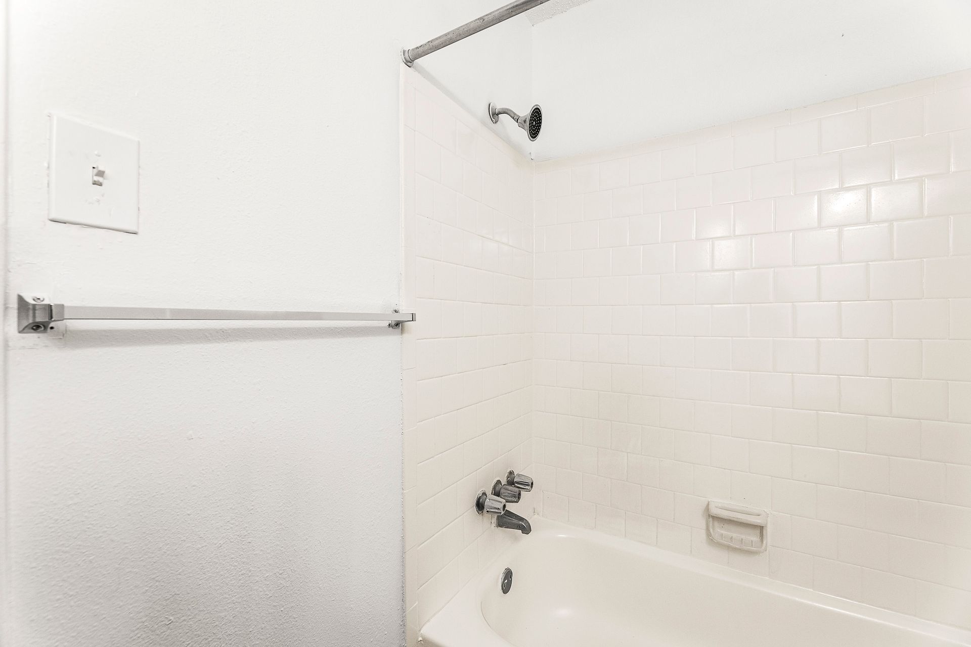 Empty white tiled bathroom with bathtub, showerhead, towel bar, and grab rail