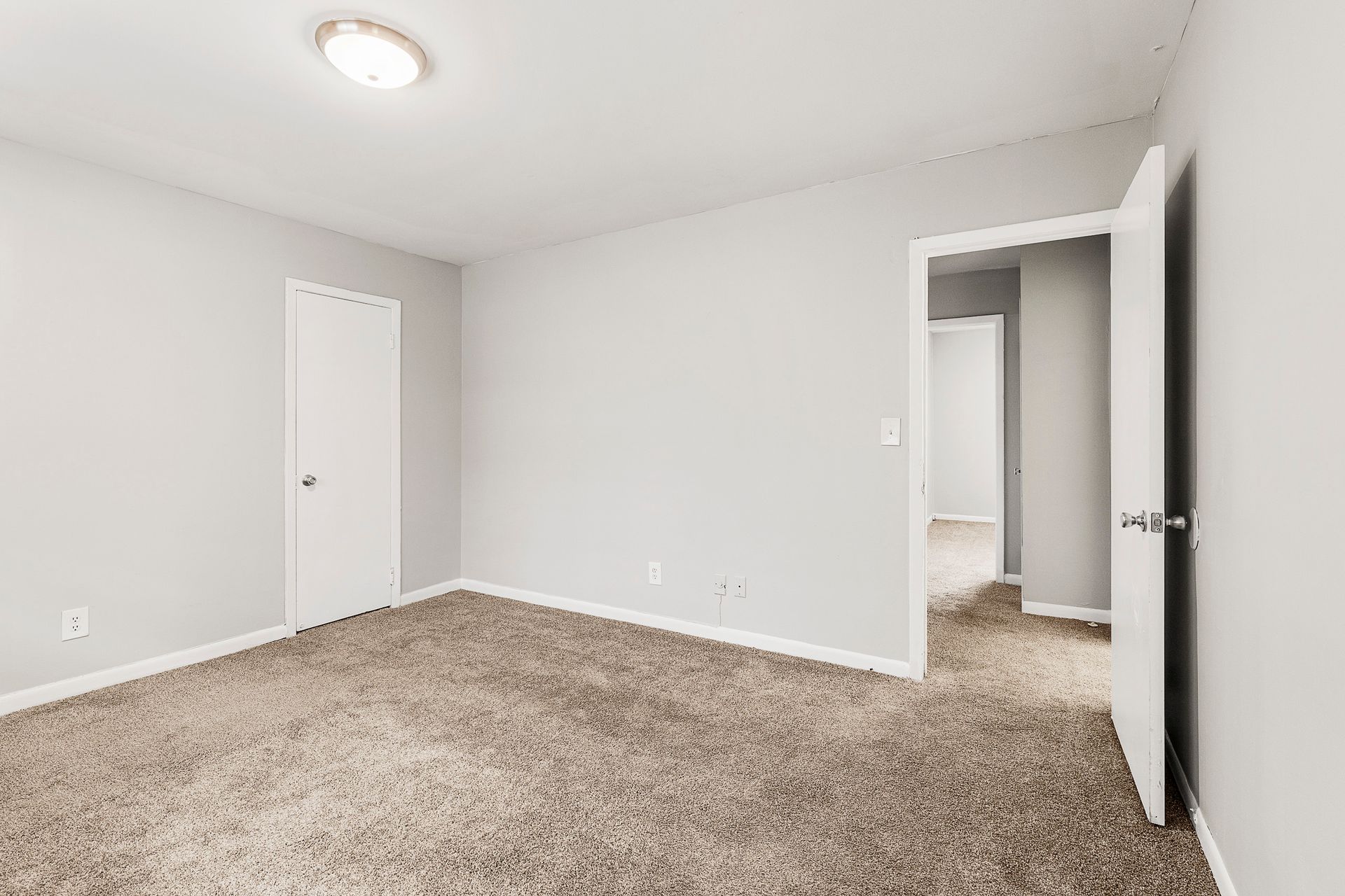 Empty beige-carpeted bedroom with light gray walls, white doors, and an open doorway.