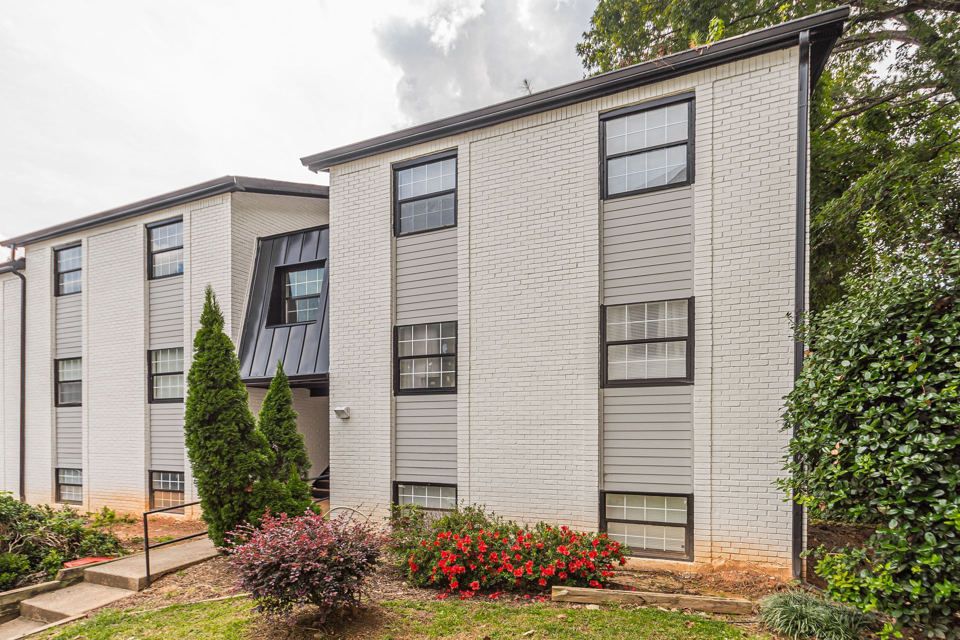 Three-story apartment building with white brick exterior, windows, and landscaped shrubs under cloudy sky