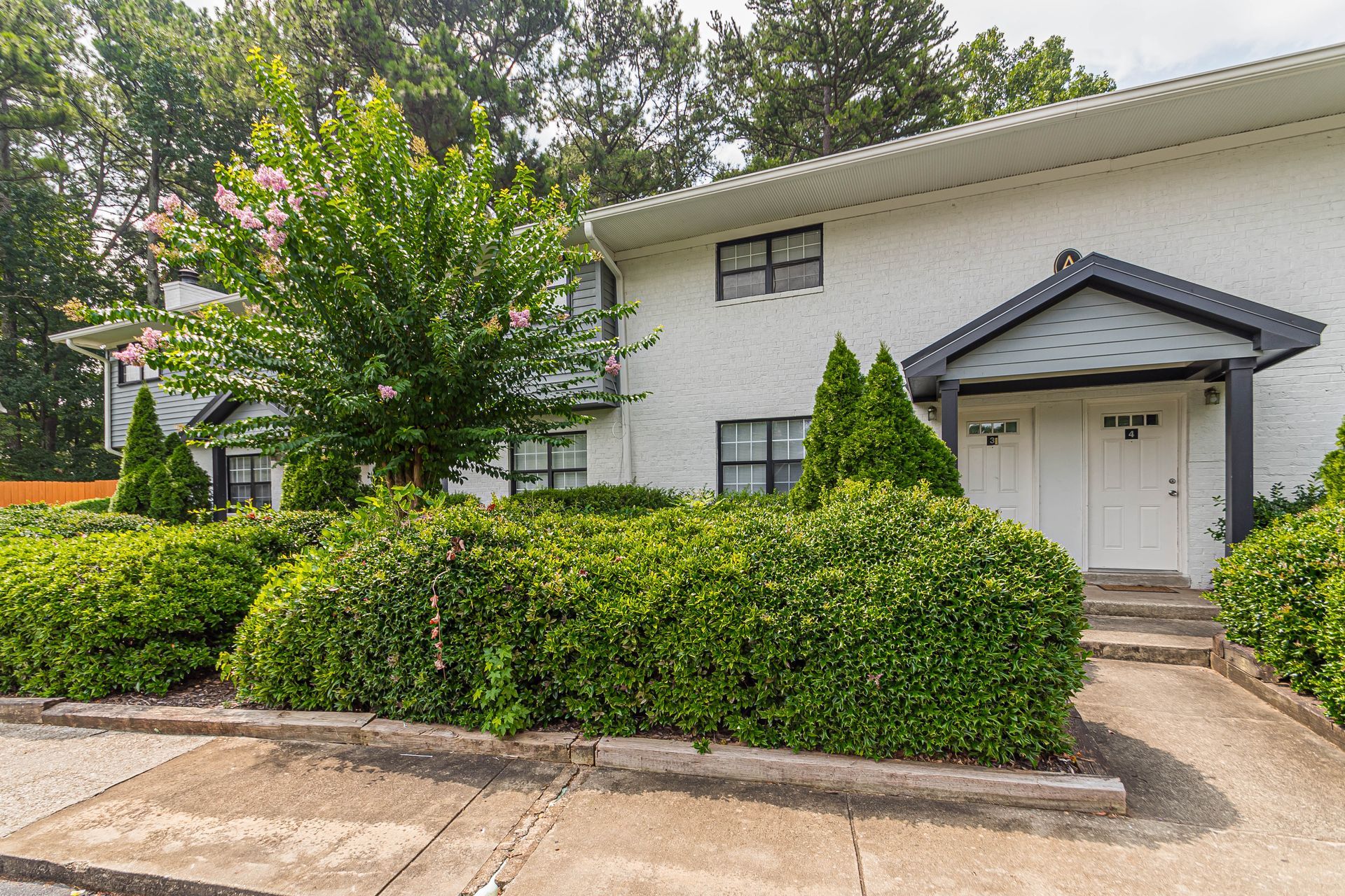 Residential apartment building with white siding, hedges, trees, and a small front porch entrance