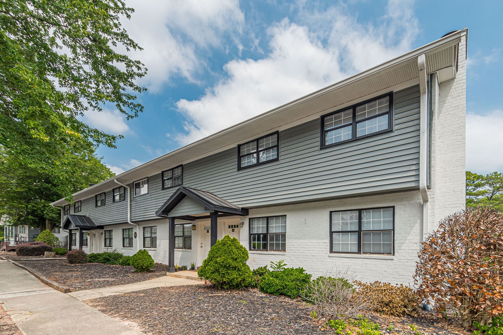 Two-story gray apartment building with white trim and a gravel parking area under a cloudy sky