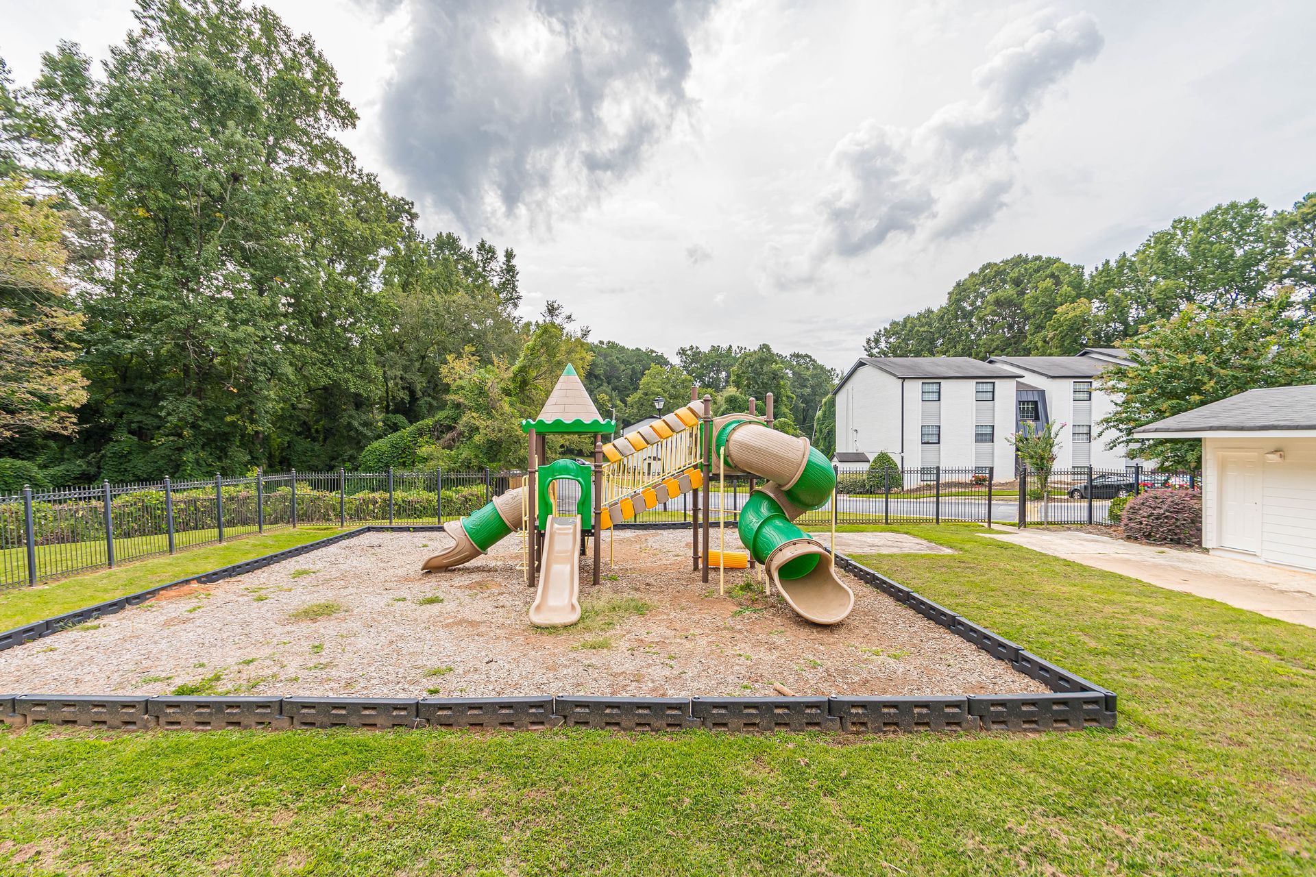 Playground with green-and-yellow slide set on a sandy area beside a grassy yard and white building