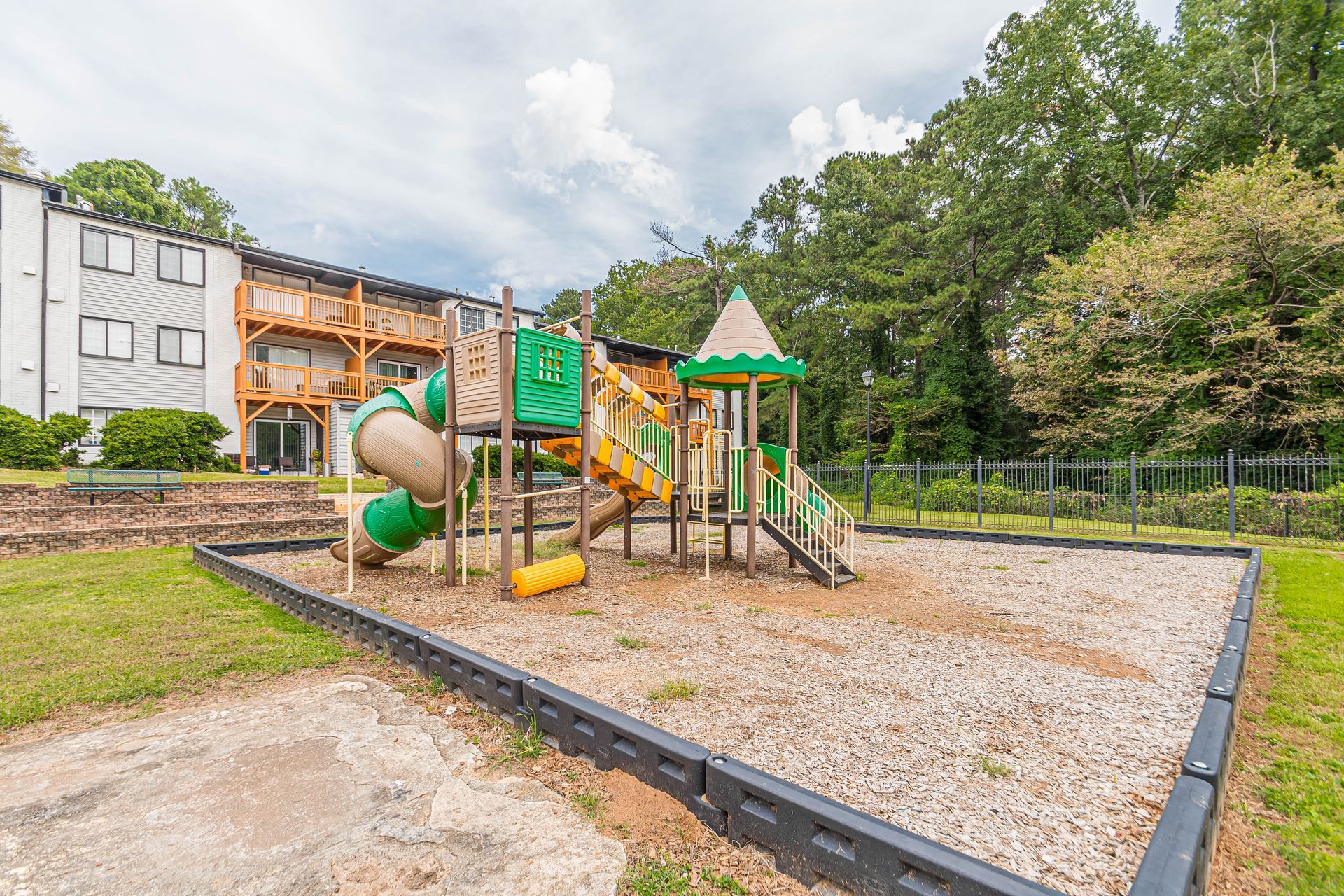 Playground with green-and-tan climbing equipment in a fenced park, bordered by gravel and grass