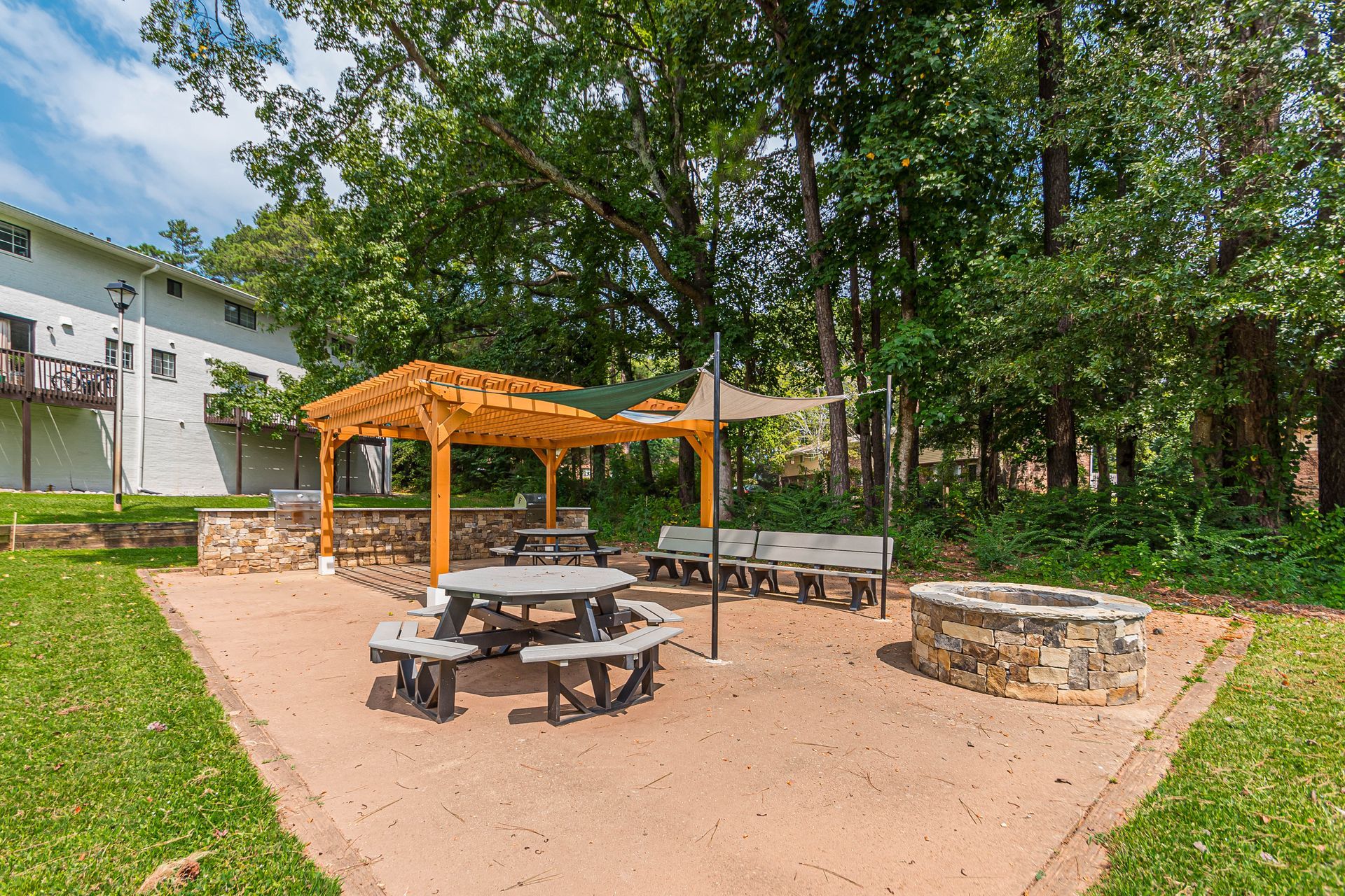 Outdoor picnic area with tables, wooden pergola, stone grill, and trees beside an apartment building