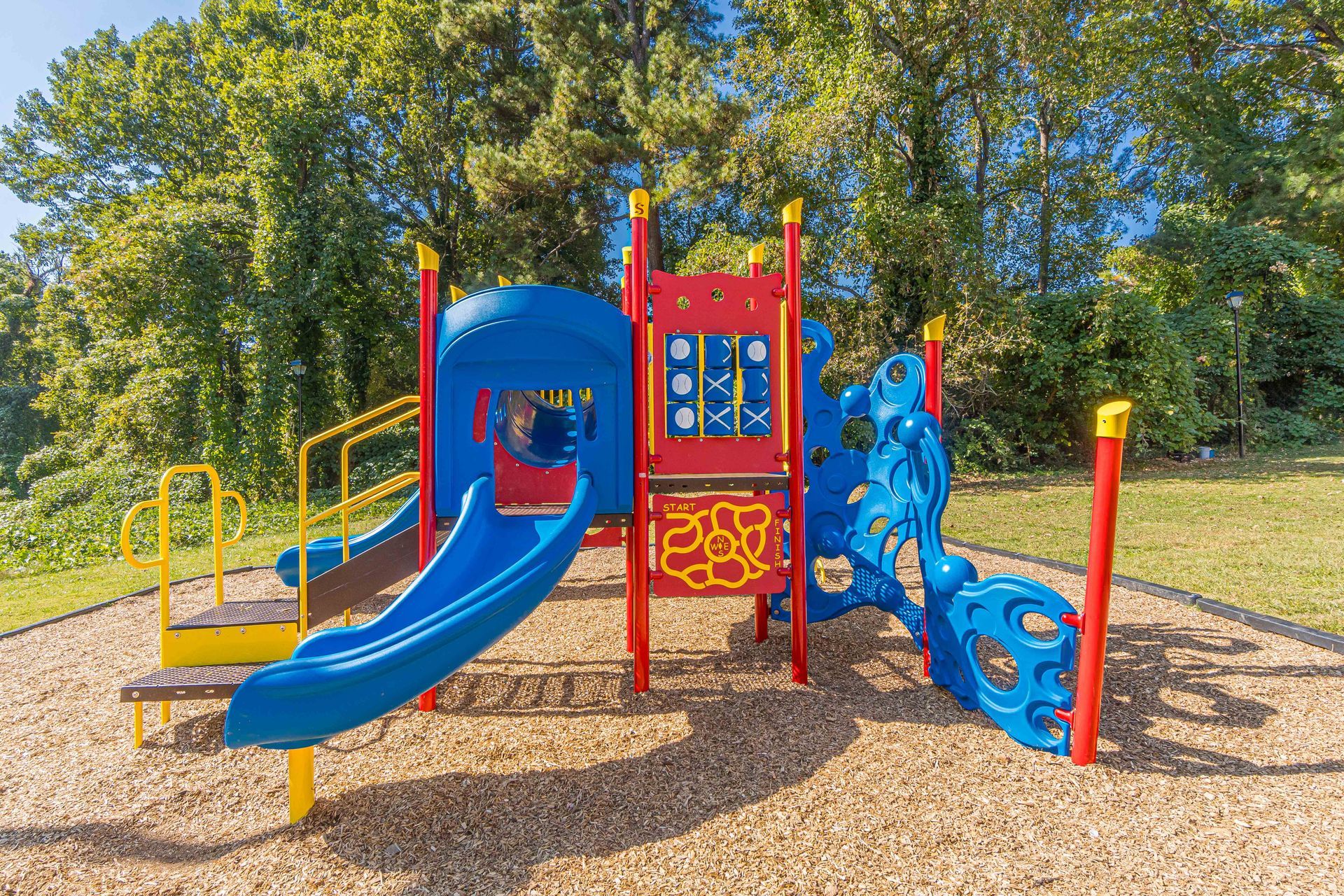 Colorful playground with blue slides, red climbing structure, and yellow rails on wood chips near trees