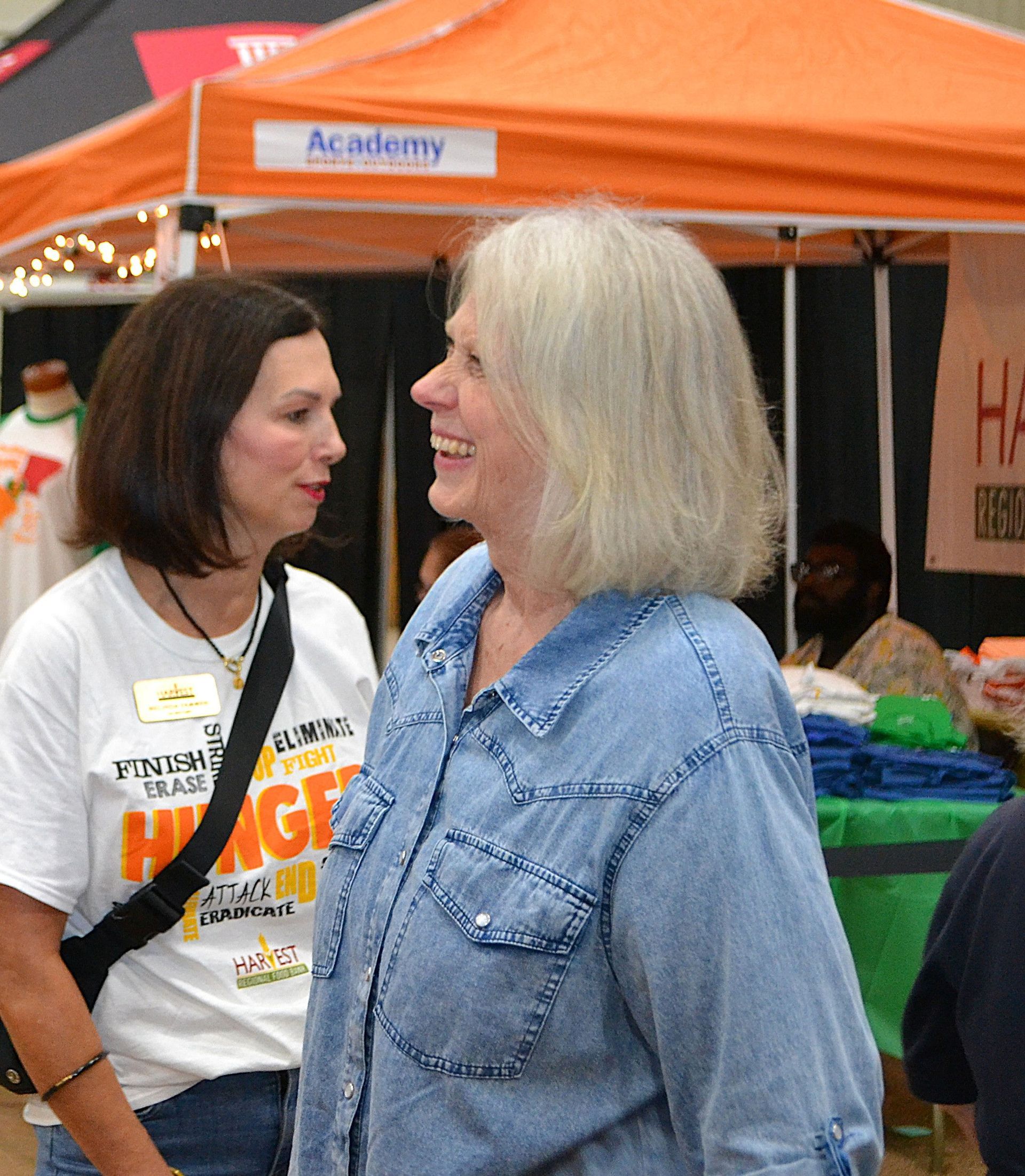 Two women chatting at an outdoor booth under an orange tent, one in a blue denim shirt.