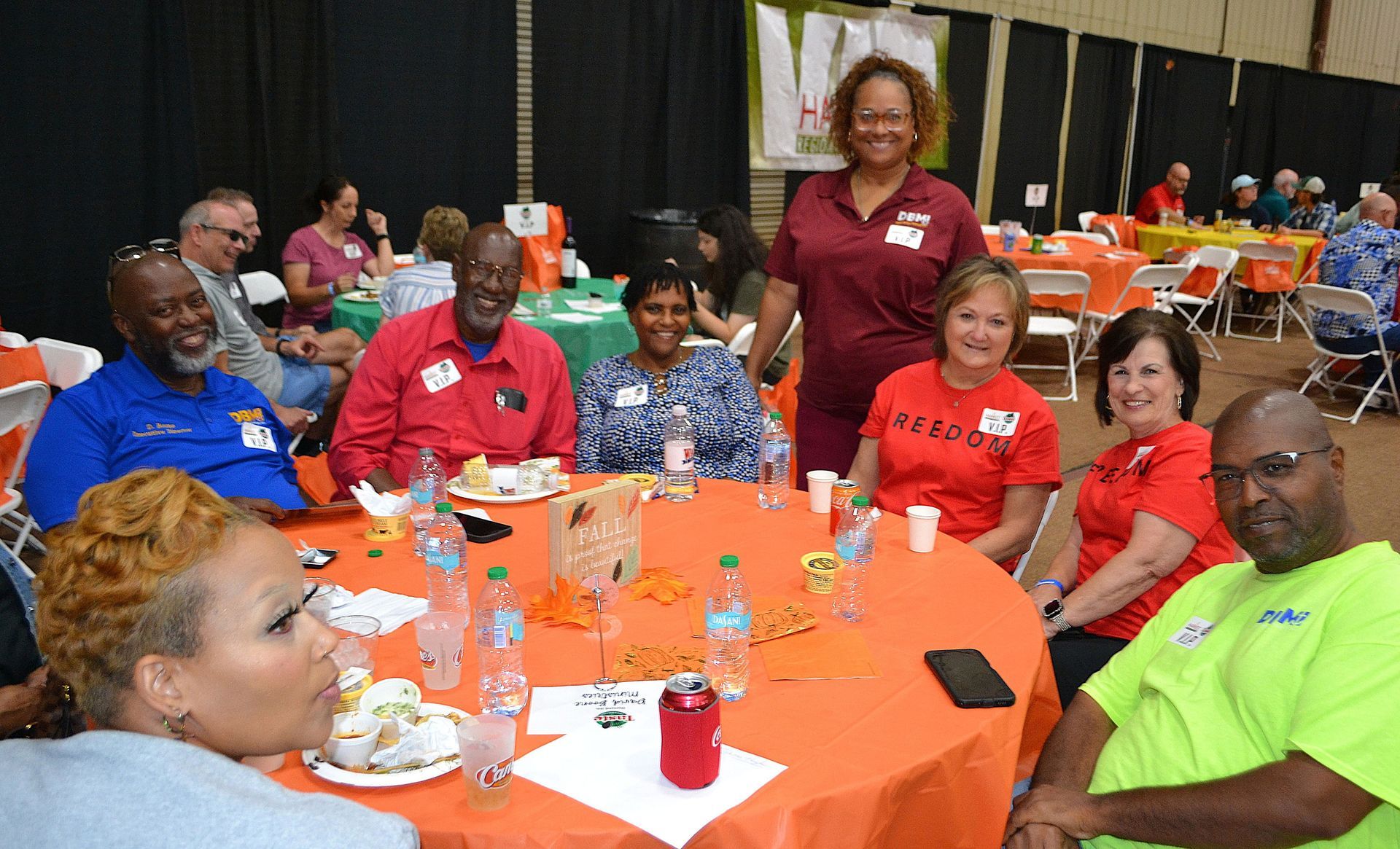 People seated around orange-clothed tables at a lively indoor gathering, smiling and posing for a group photo