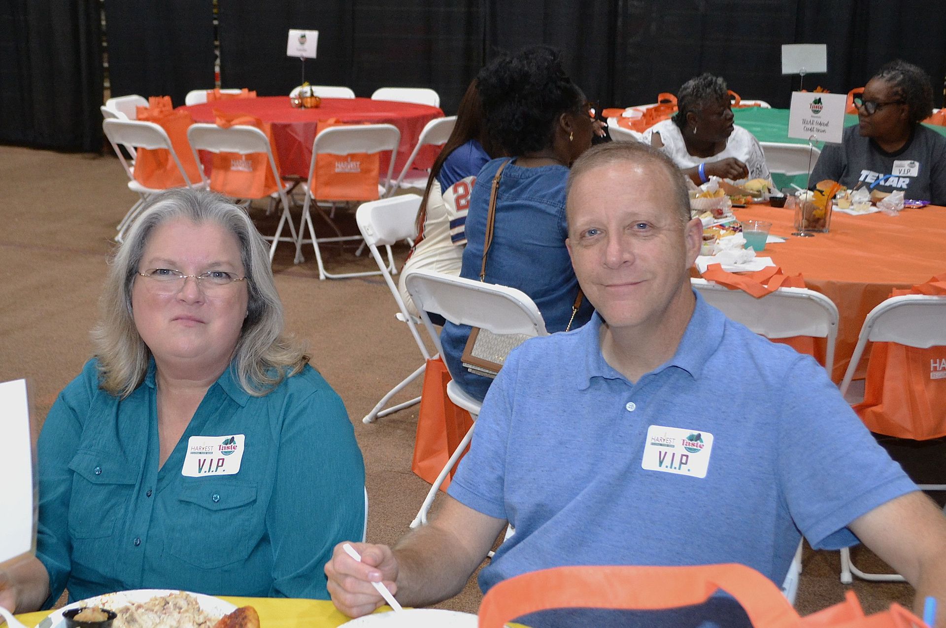 Two people seated at a banquet table in a hall with orange-covered chairs and round tables