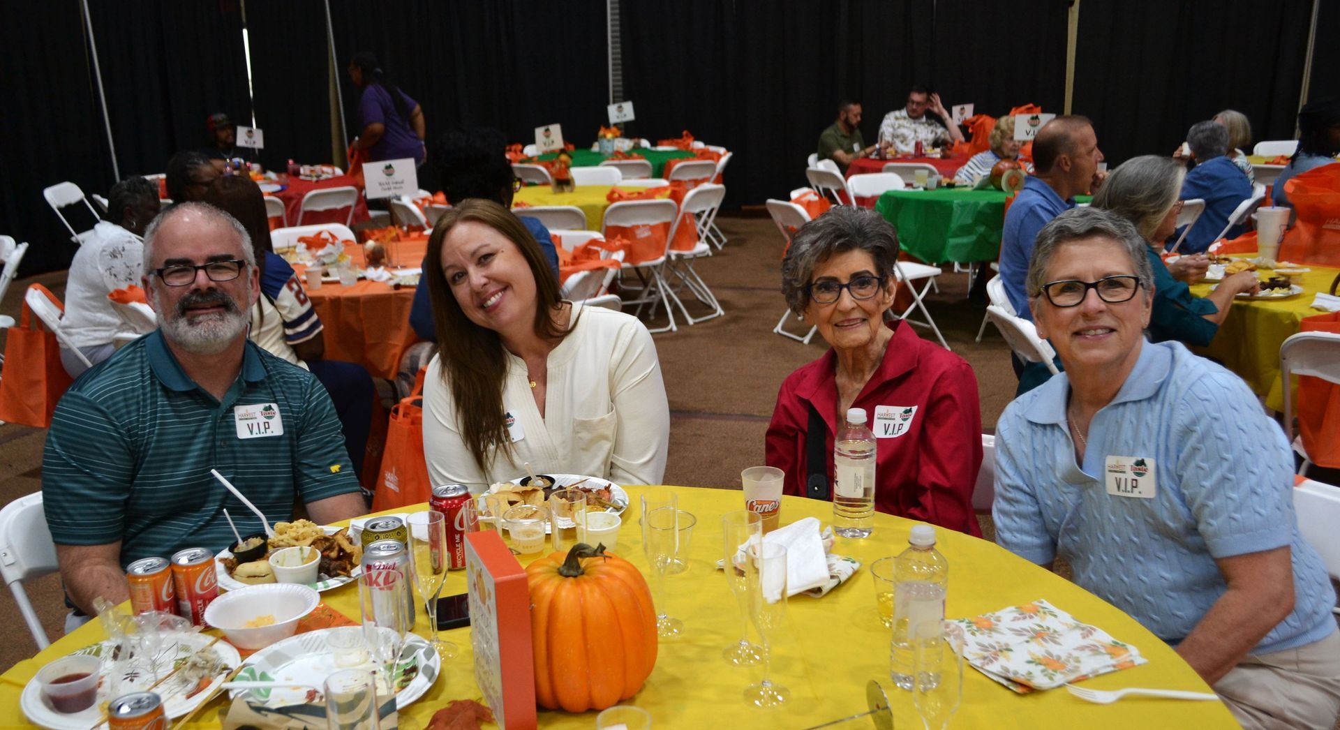 Four people seated at a yellow table with drinks and a pumpkin centerpiece at a festive indoor event