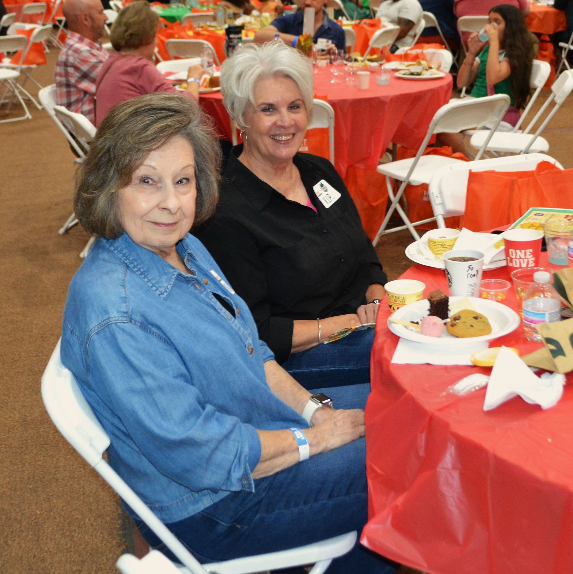 Two women seated at a red-covered banquet table, smiling with plates of food in a crowded hall