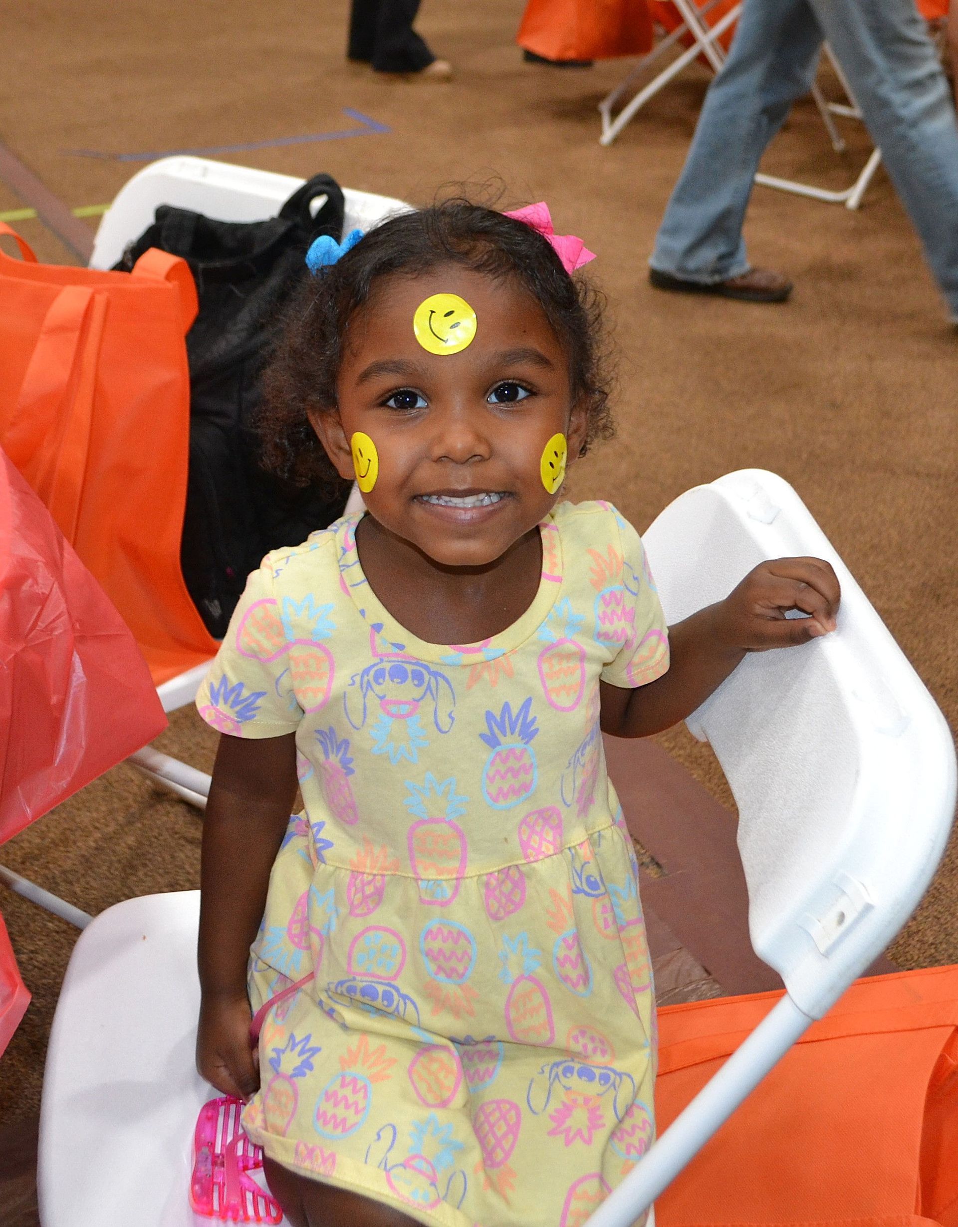 Smiling child with face paint sitting on a white chair indoors, wearing a pastel dress.