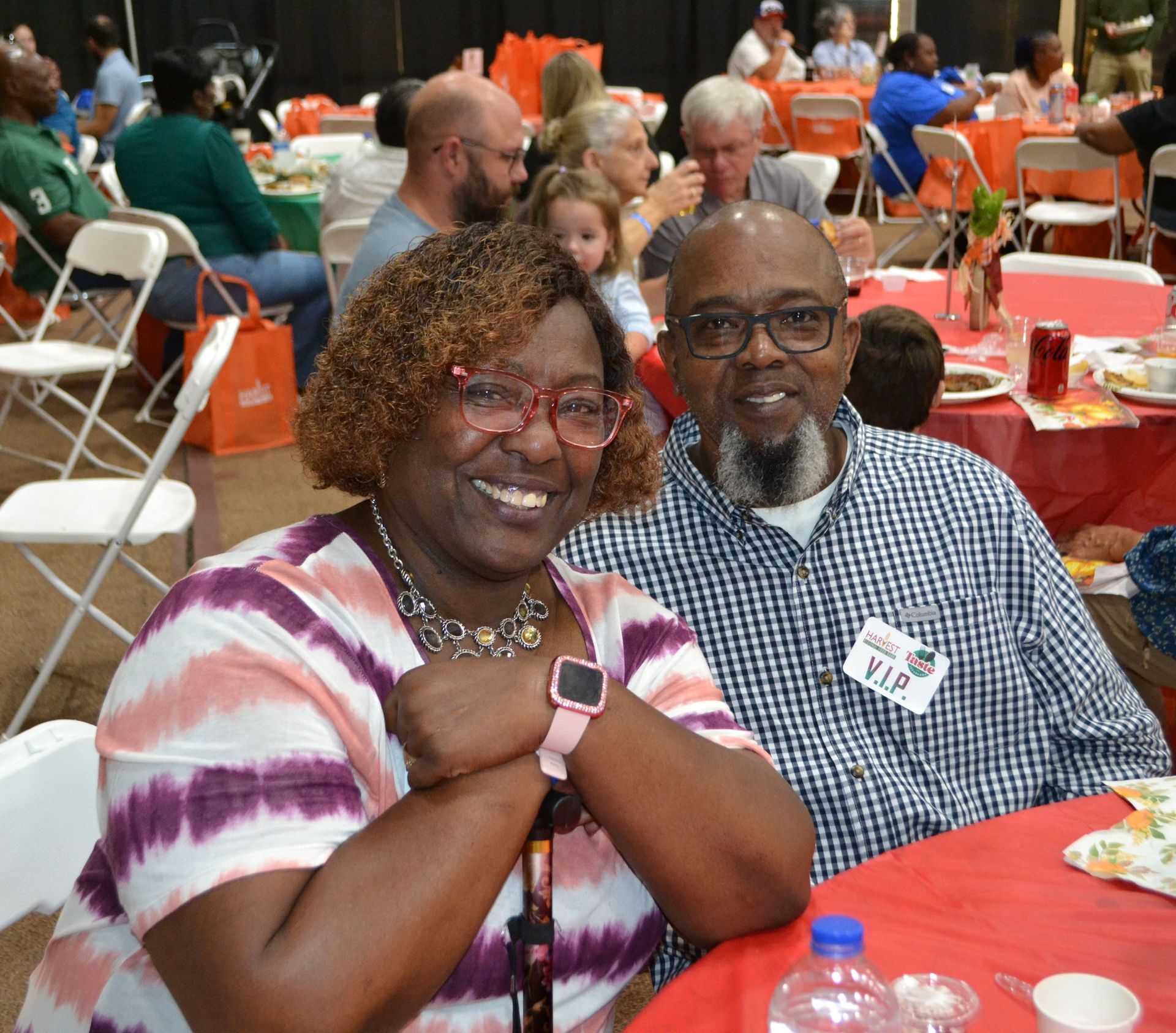 Smiling couple seated at a banquet table with red tablecloths in a crowded event hall.