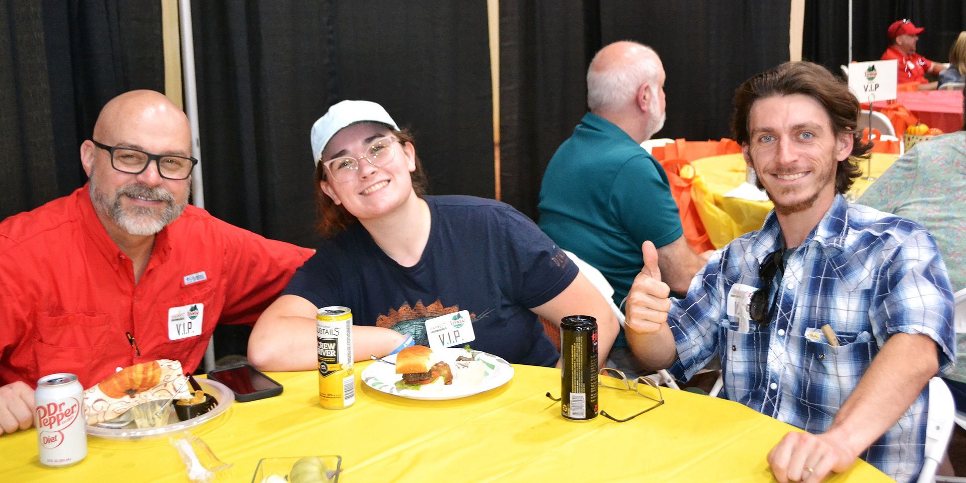 Three people seated at a yellow table, smiling at a casual indoor gathering with food and drinks.