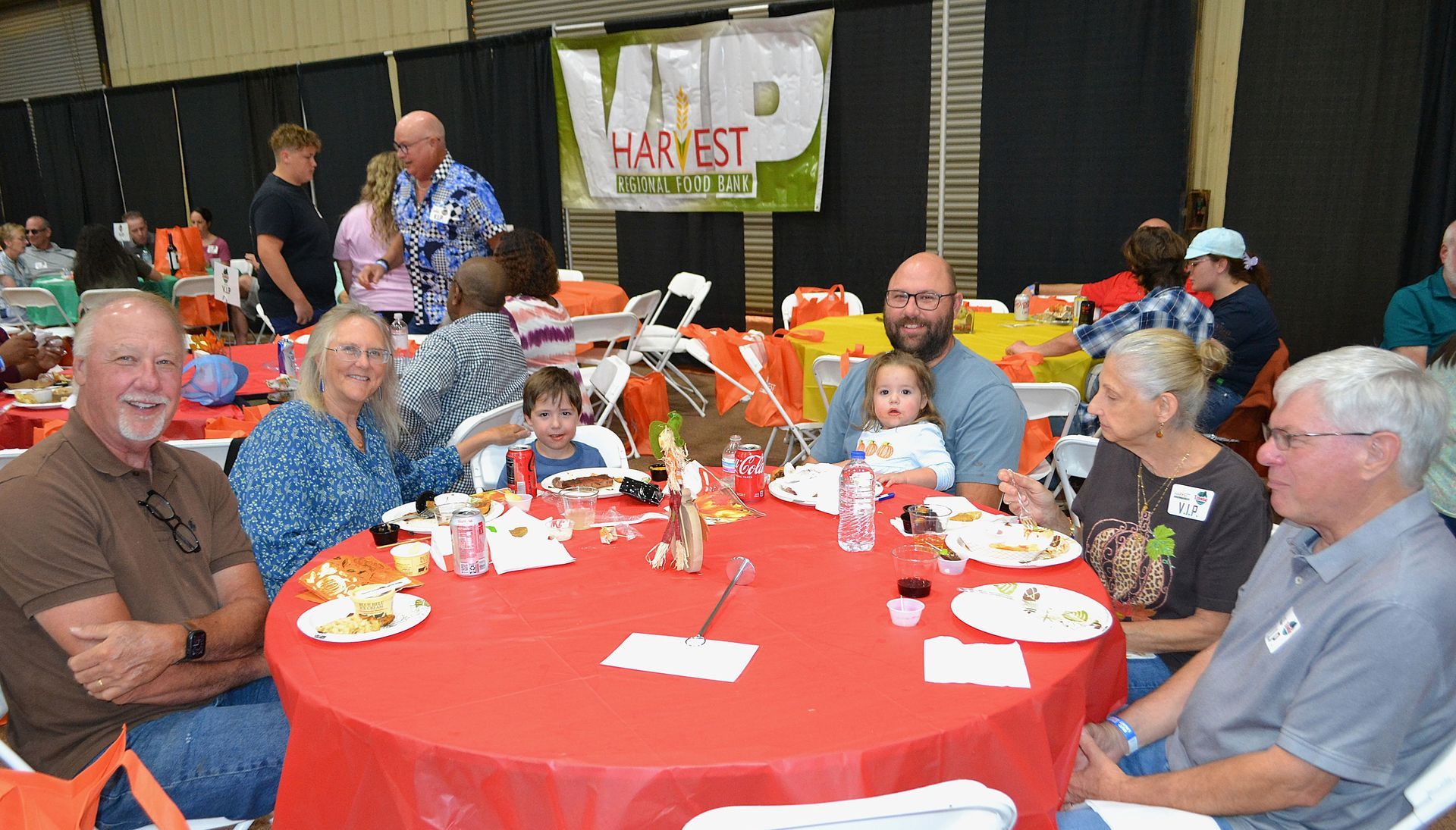 People seated at a red-tablecloth banquet table in a festive hall, with balloons and a banner behind them