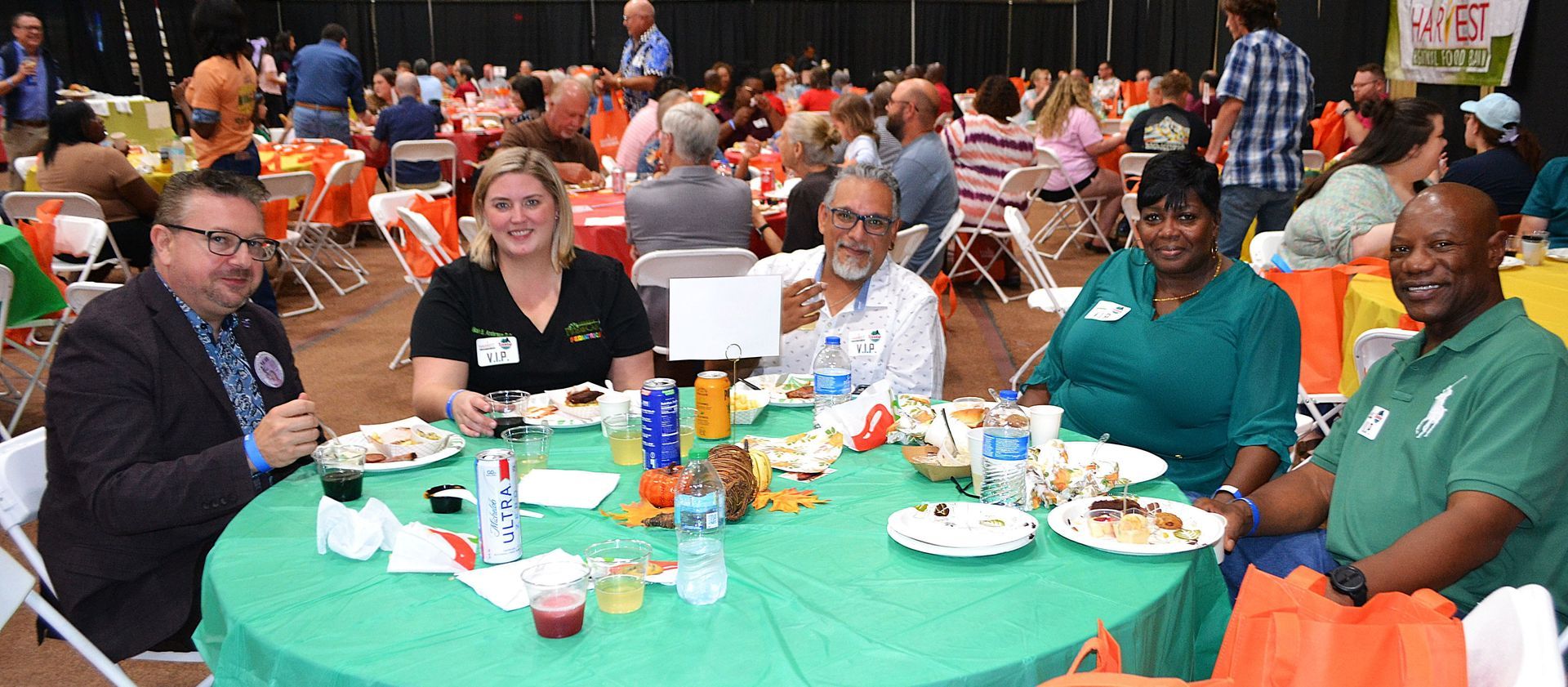 People seated at a banquet table with food and drinks in a busy indoor event hall