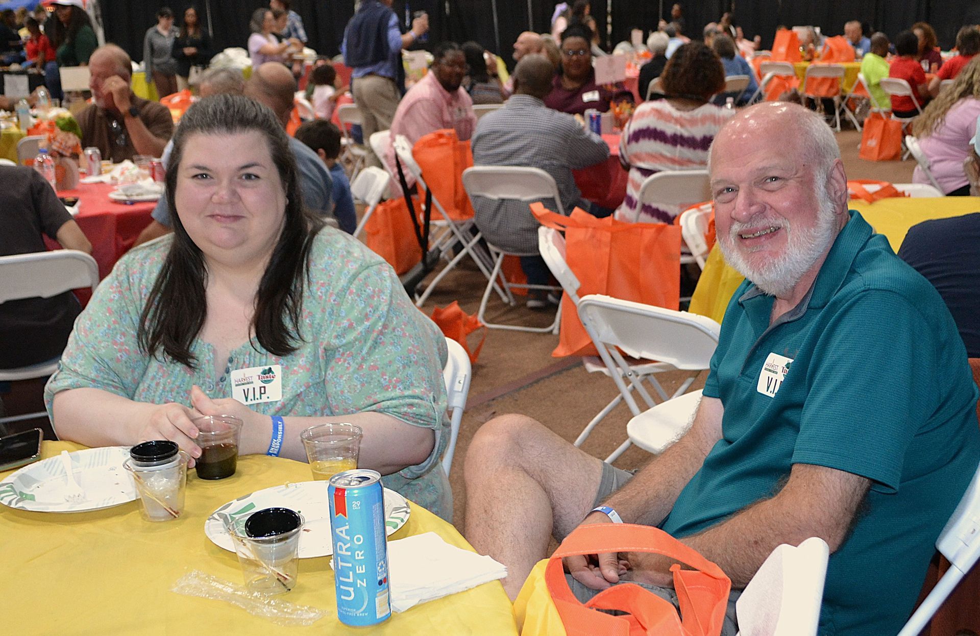 Two people seated at a banquet table with food and drinks in a busy hall