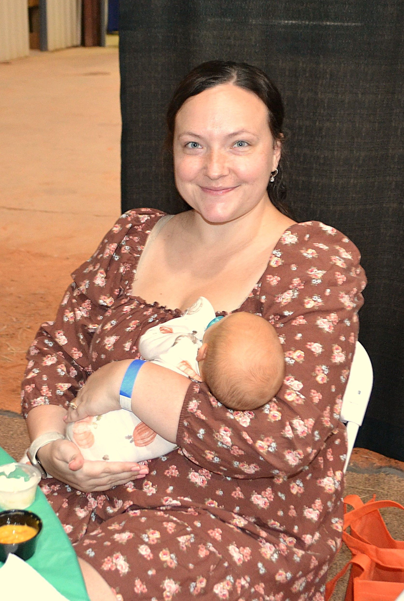 Woman holding two babies, seated indoors and smiling in a brown patterned dress.