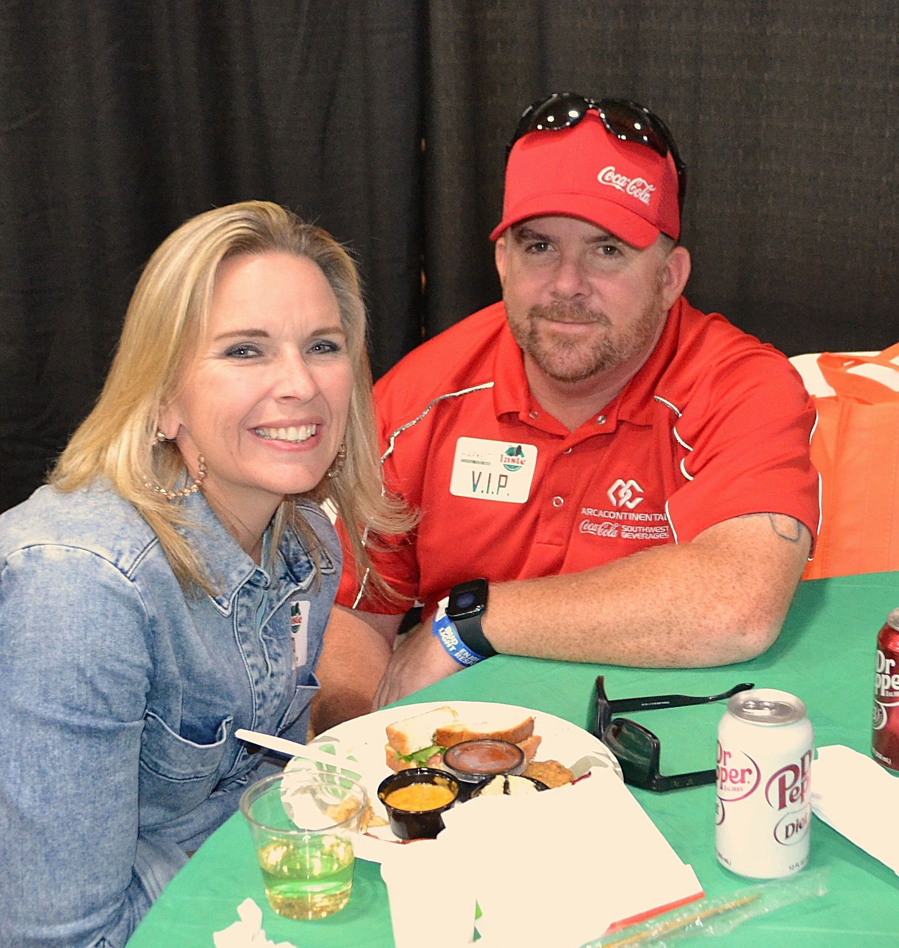 Two people seated at a table with food and drinks, smiling at an indoor event