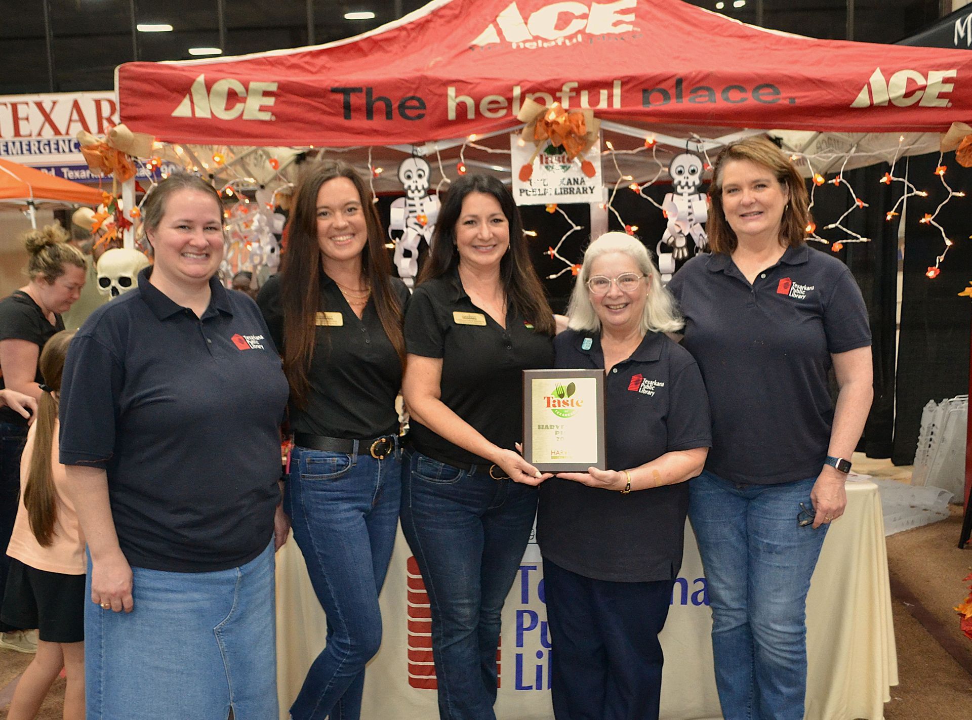 Five people posing at an ACE event booth, holding a framed award, with red ACE banners behind them