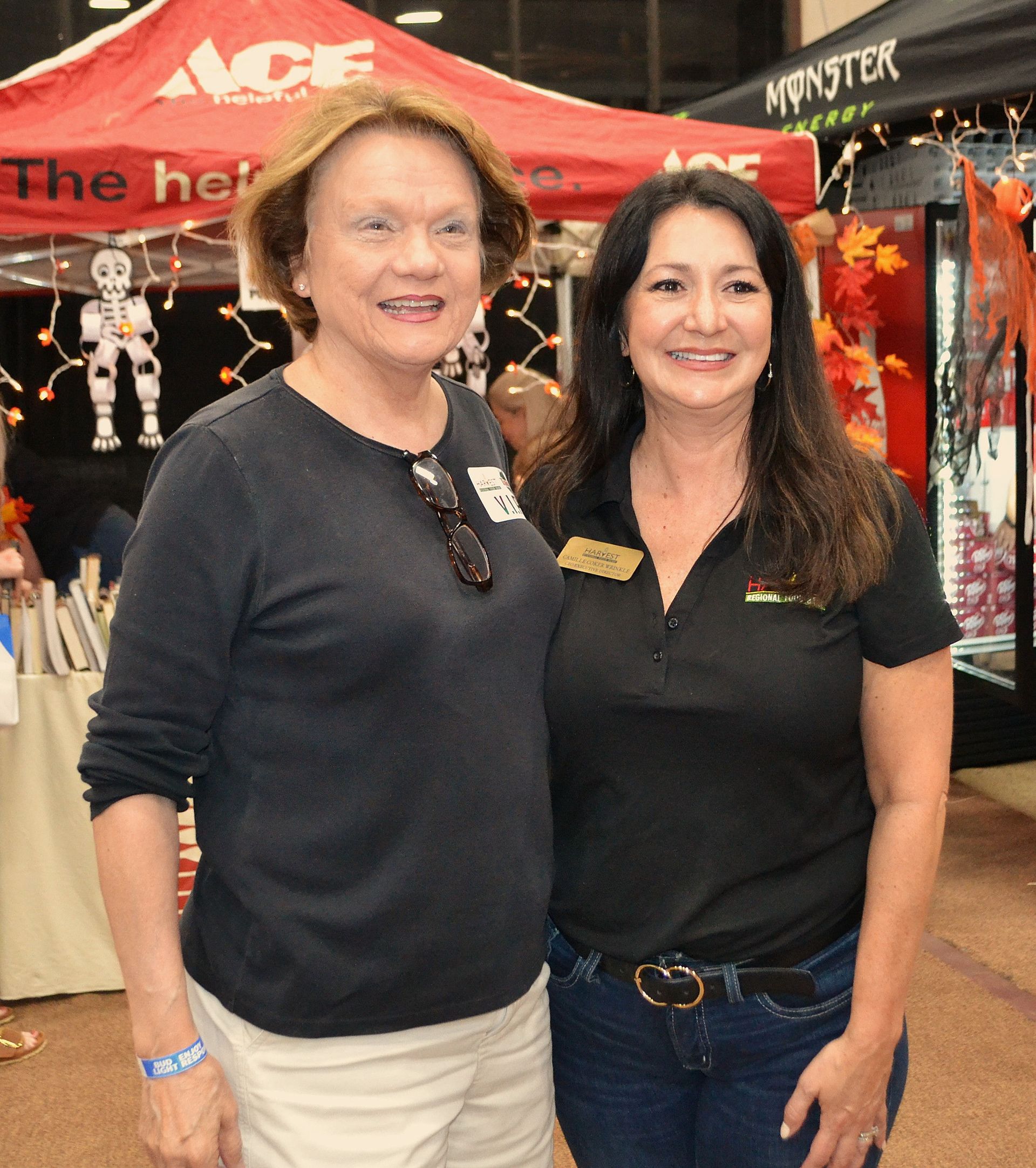 Two smiling women stand at an indoor booth with red tents and a Monster sign behind them.