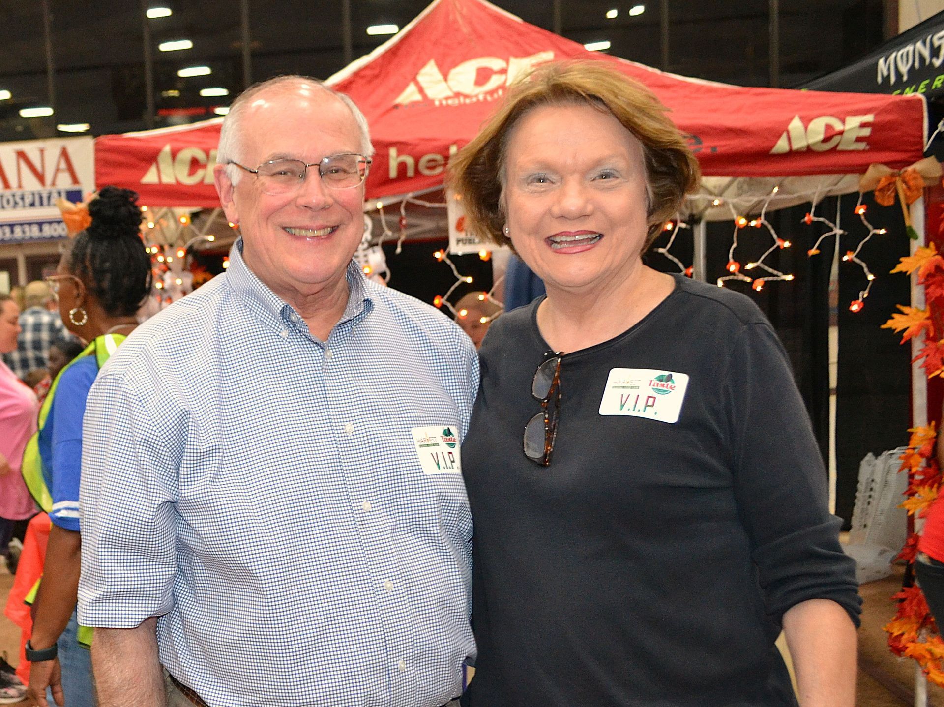 Two smiling people standing in front of a red ACE canopy at a market booth
