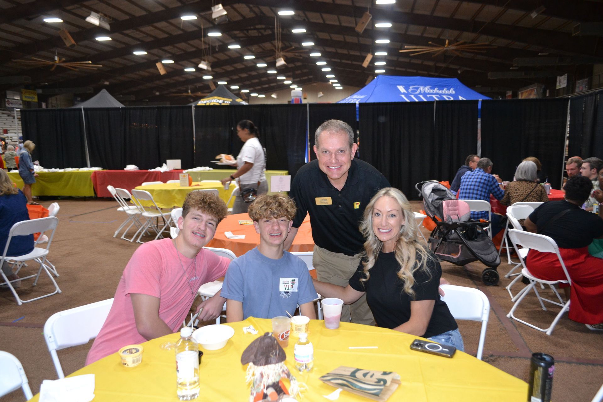 People seated at a yellow table in a large indoor event hall, smiling toward the camera.