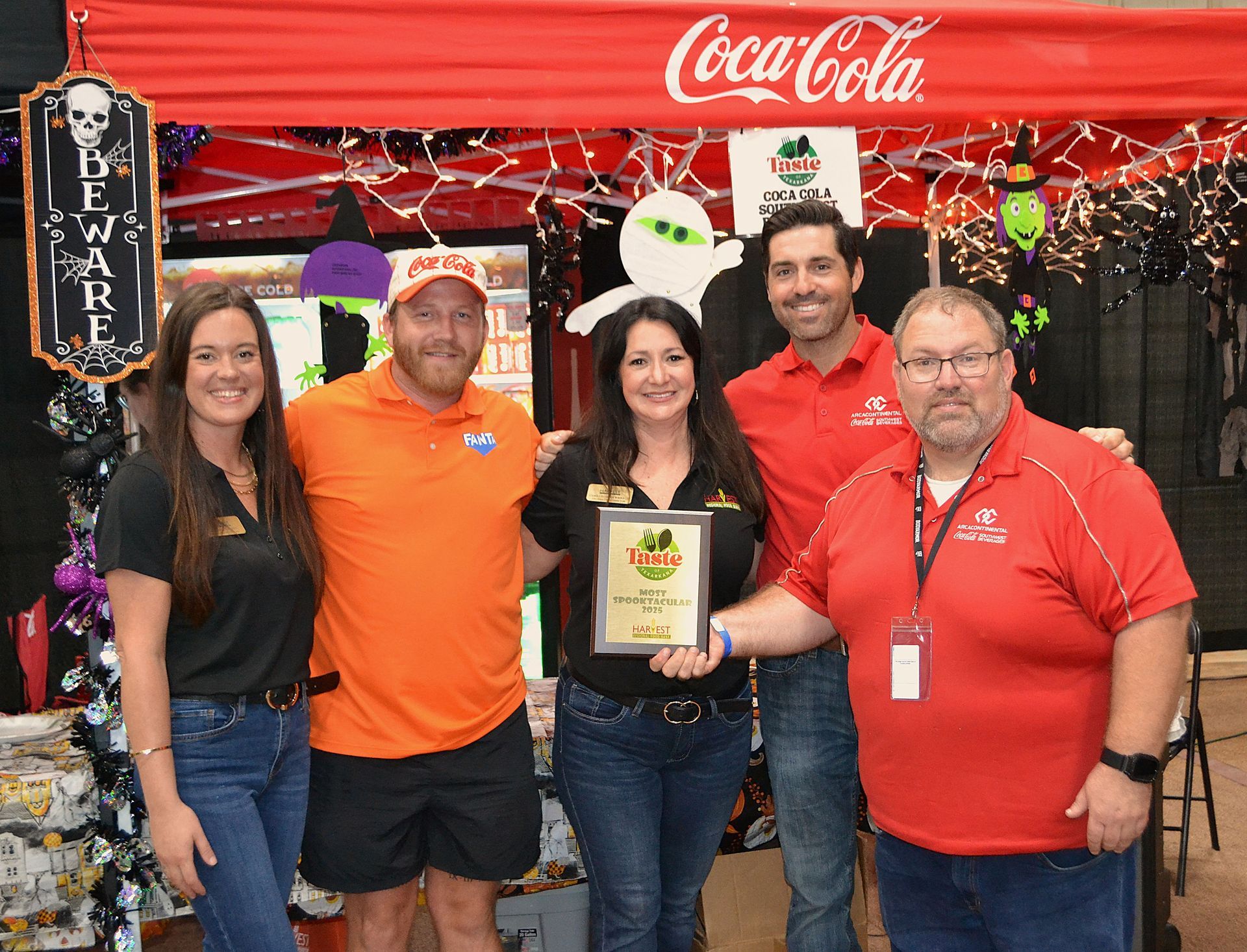 Five people posing at a Coca-Cola booth, holding a framed award in a festive red-decorated setting.