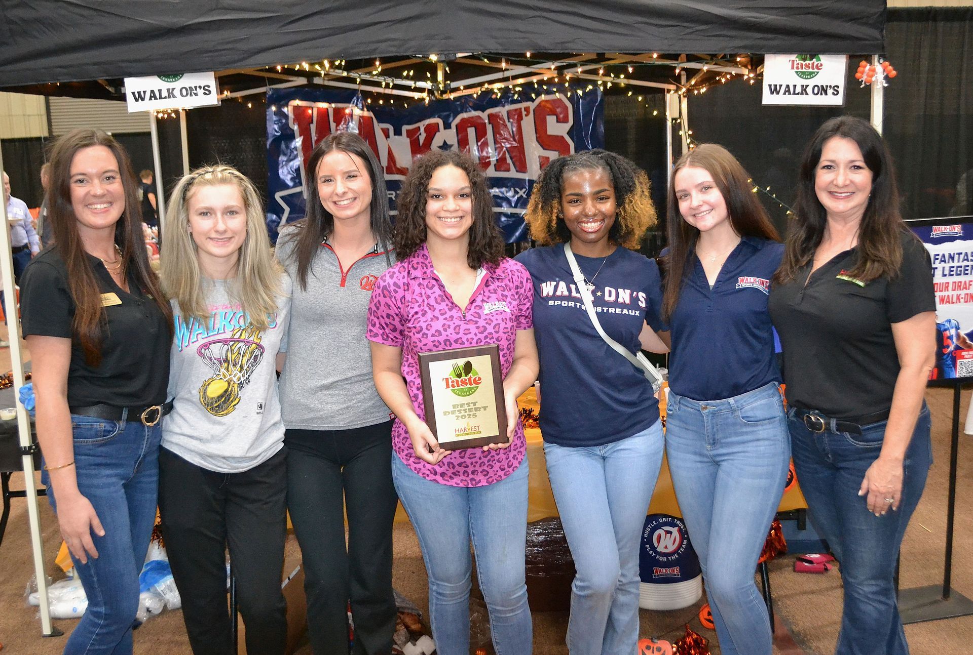 Seven people standing at a fair booth, smiling and holding a sign under a canopy.