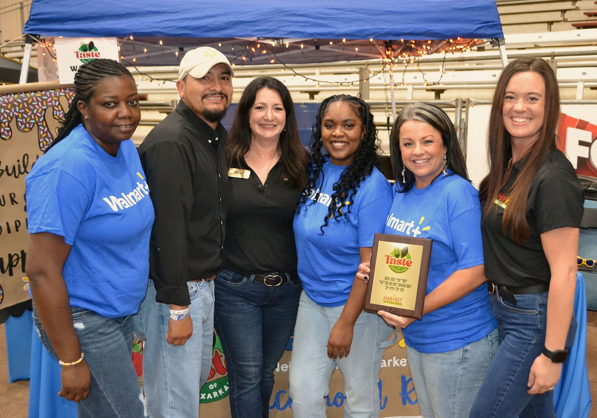 Five people standing together at an outdoor event booth, smiling and holding a framed plaque.