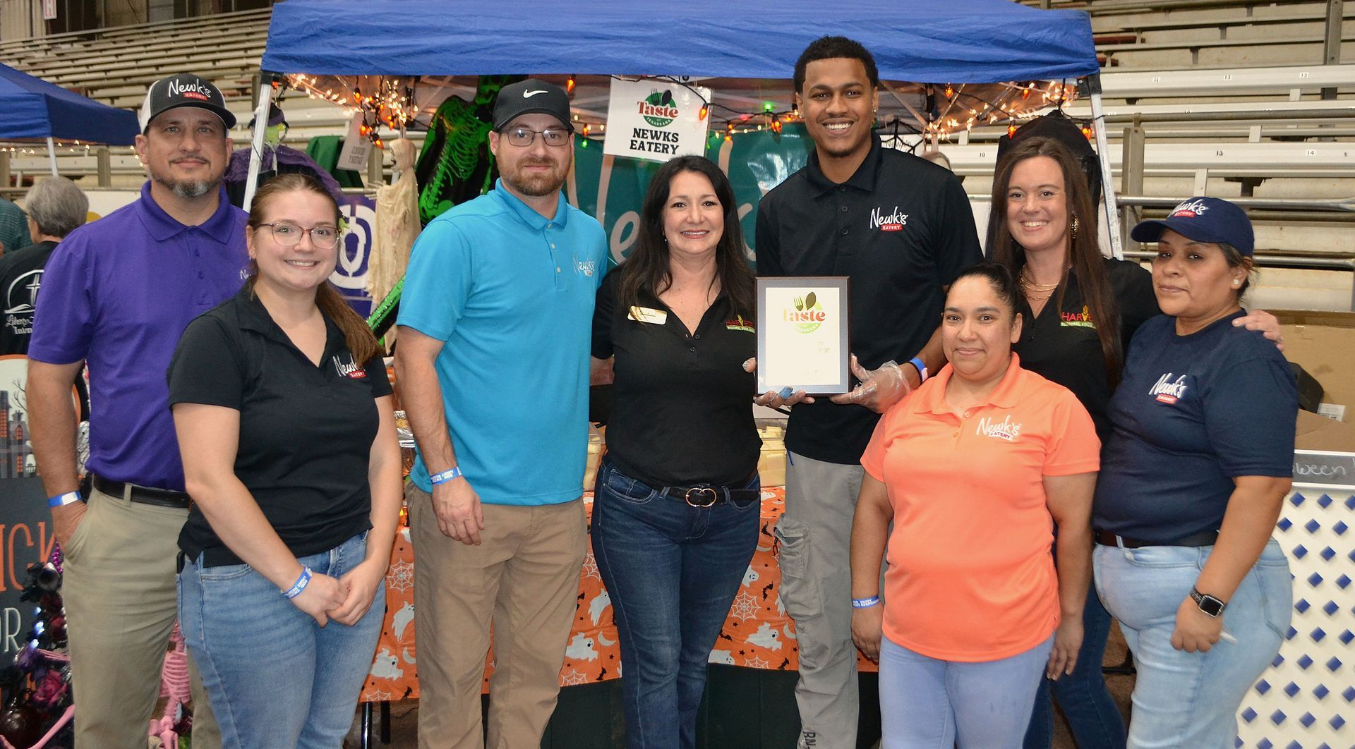 Group of people standing at an outdoor market booth, posing with a plaque and smiling under a blue canopy.